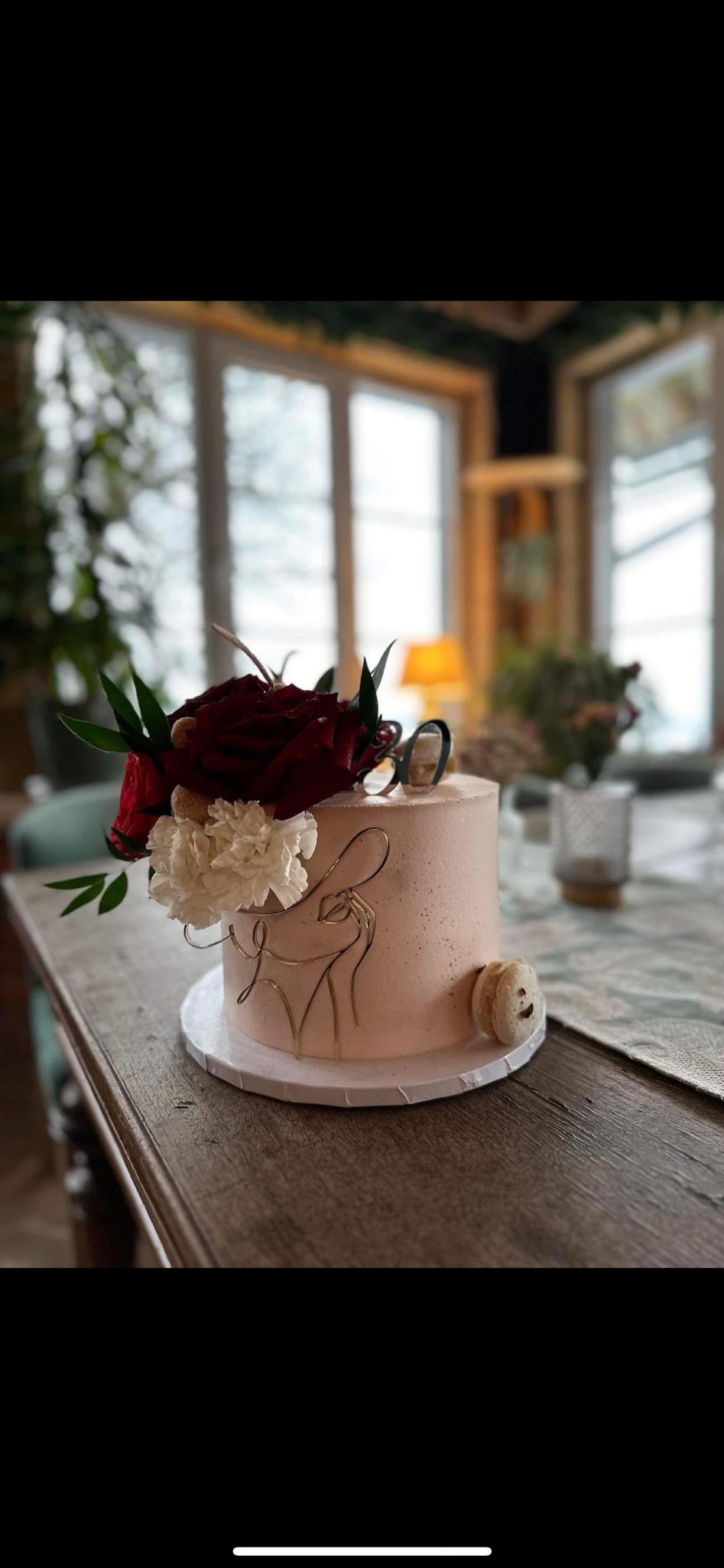 Tort urodzinowy w pudrowym różu, ozdobiony kompozycją z ciemnoczerwonych i białych kwiatów, złotym konturem kobiety i uśmiechniętym makaronikiem, na drewnianym stole w jasnym wnętrzu.