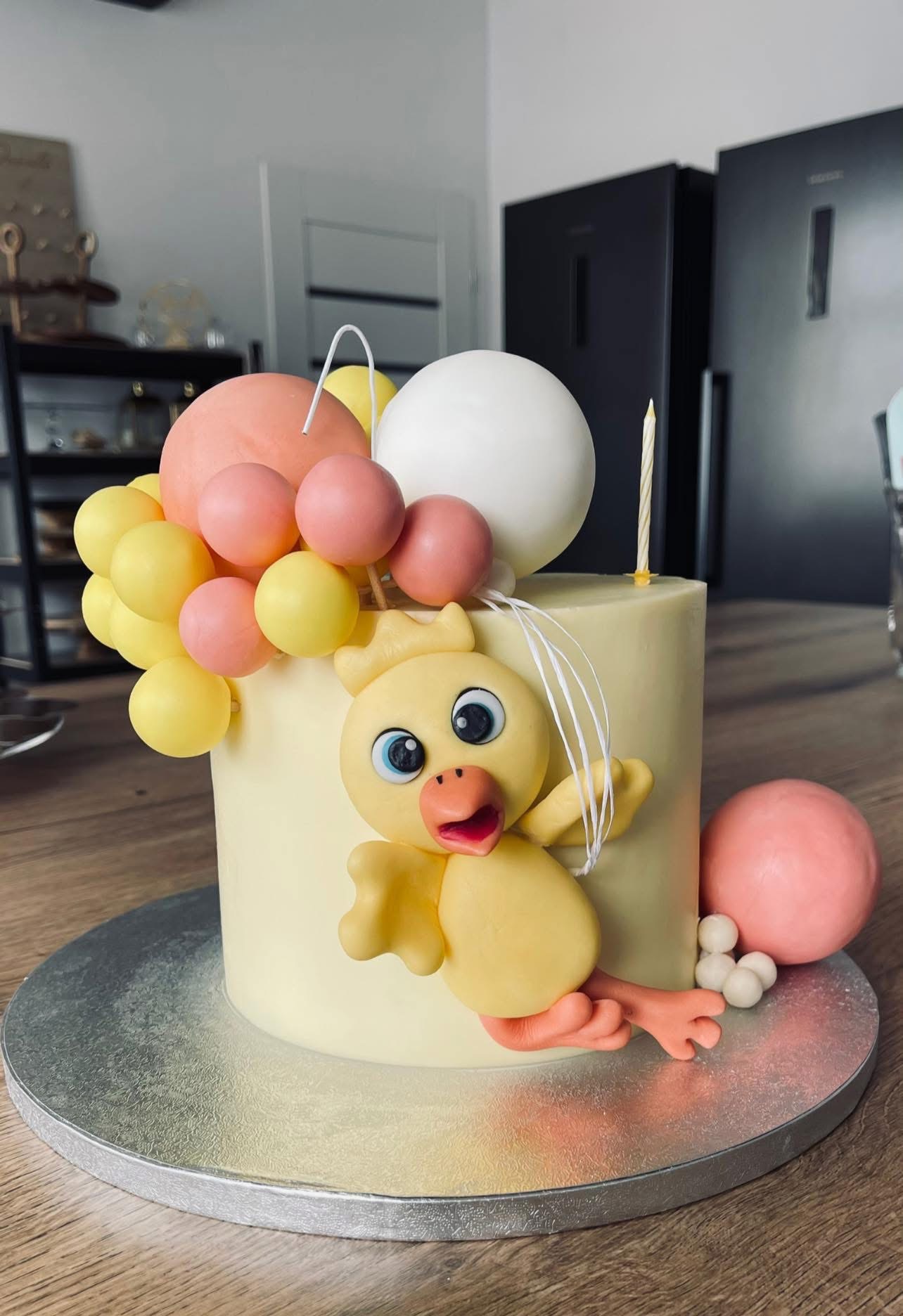 Tort urodzinowy z figurką kurczaka z modeliny, otoczony balonami z lukru, z zapaloną świeczką na tle nowoczesnej kuchni.