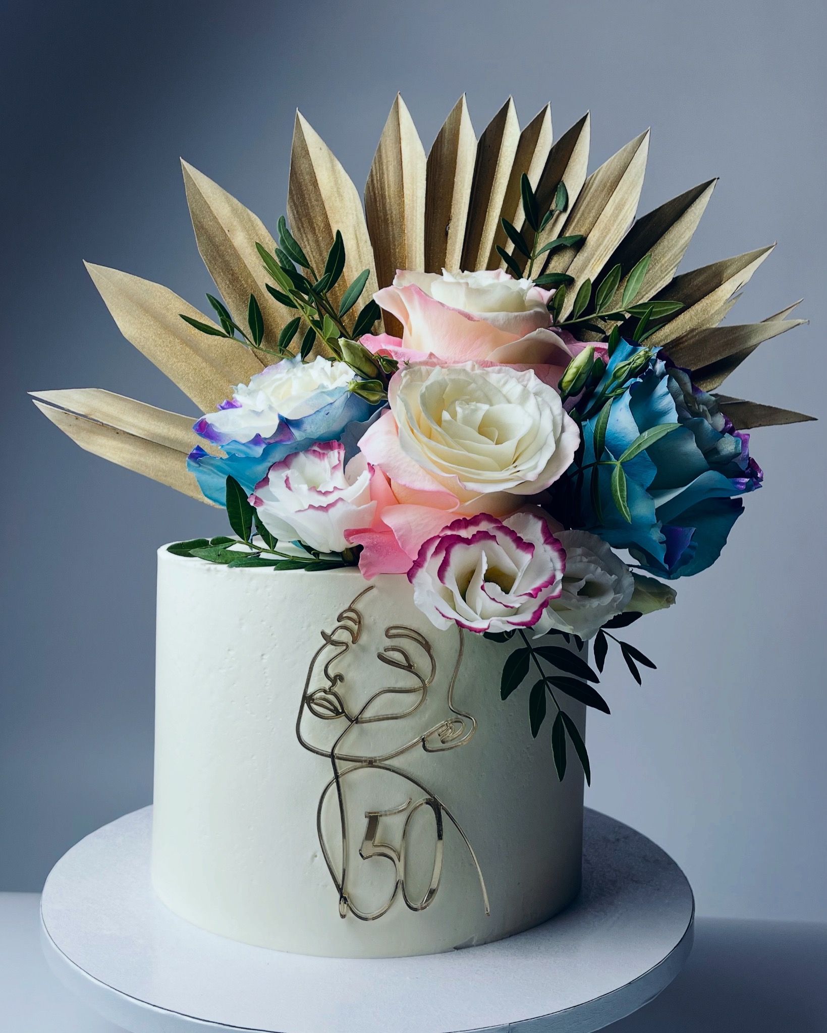 Tort urodzinowy z białym kremem, dekoracją z kwiatów w odcieniach bieli, różu i błękitu, złotymi liśćmi palmowymi oraz konturem twarzy i cyfrą 50 wykonanymi z drutu.
