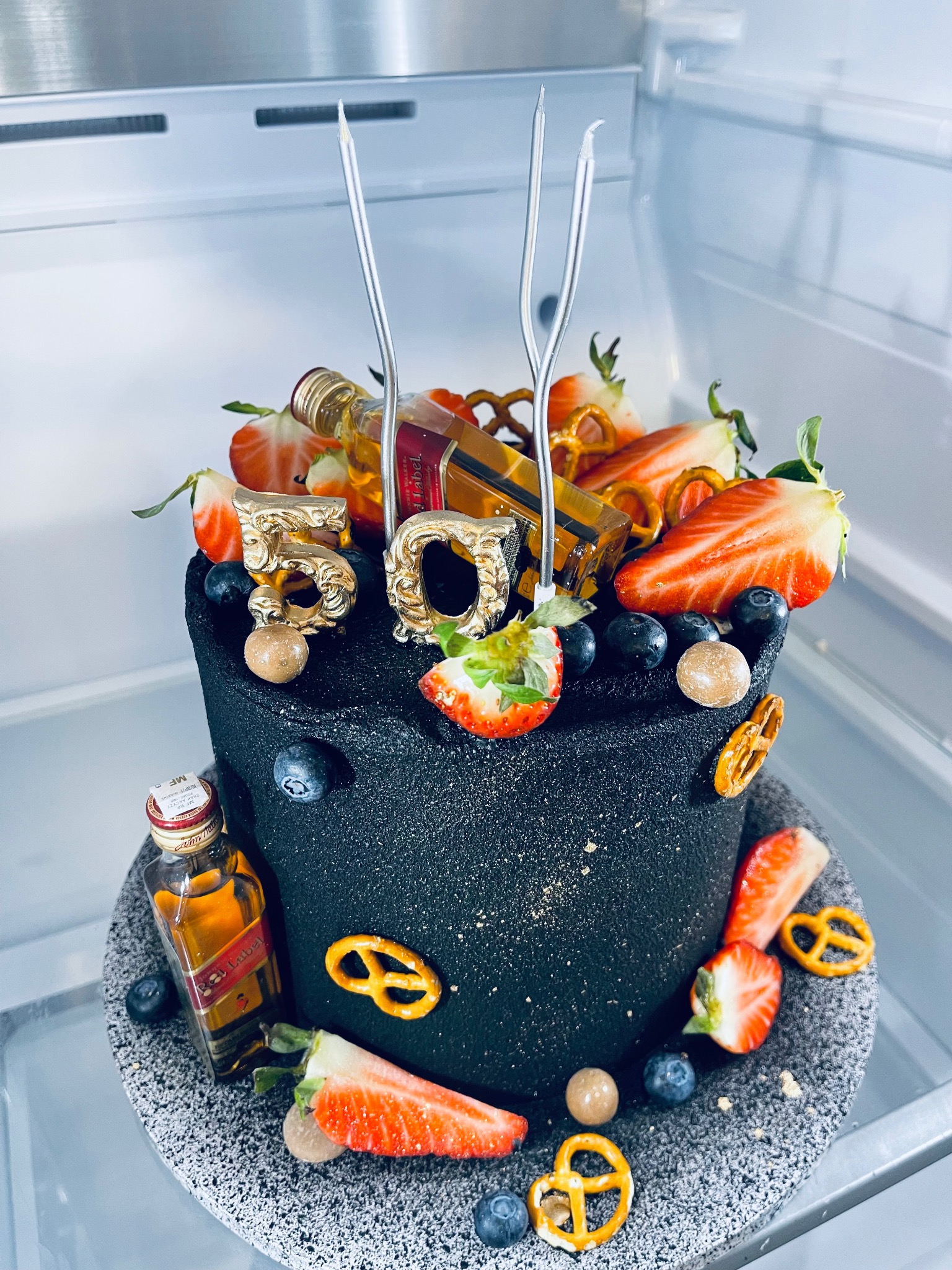 Czarny tort urodzinowy z dekoracjami z truskawek, borówek, precelków, złotych cyfr '50' i miniaturowych butelek alkoholu, umieszczony na szarym talerzu w lodówce.