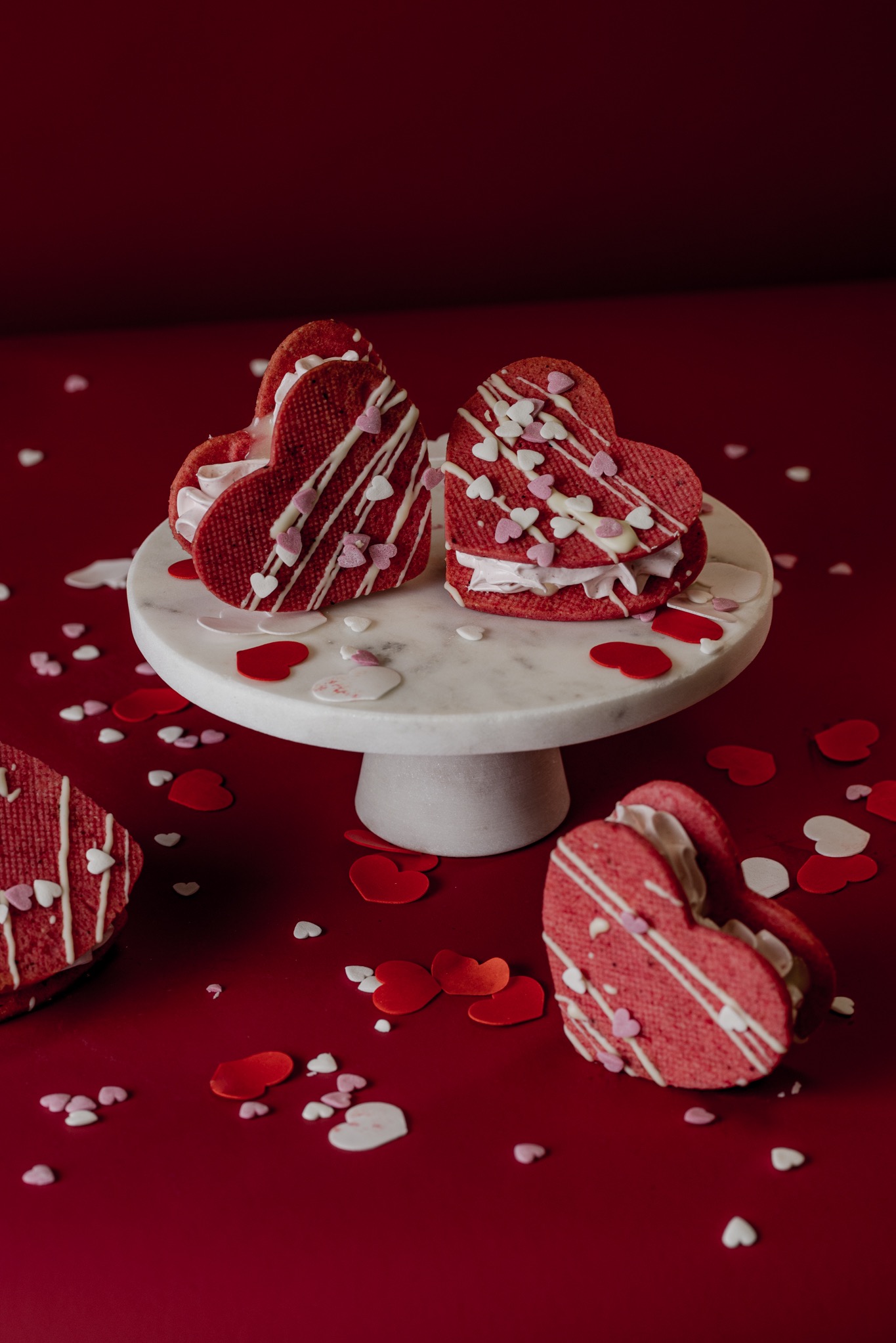 Walentynkowe ciasteczka w kształcie serc przełożone kremem, udekorowane lukrem i posypką w kształcie serduszek, ustawione na marmurowym stojaku na tle czerwonego tła z rozsypanymi dekoracjami.