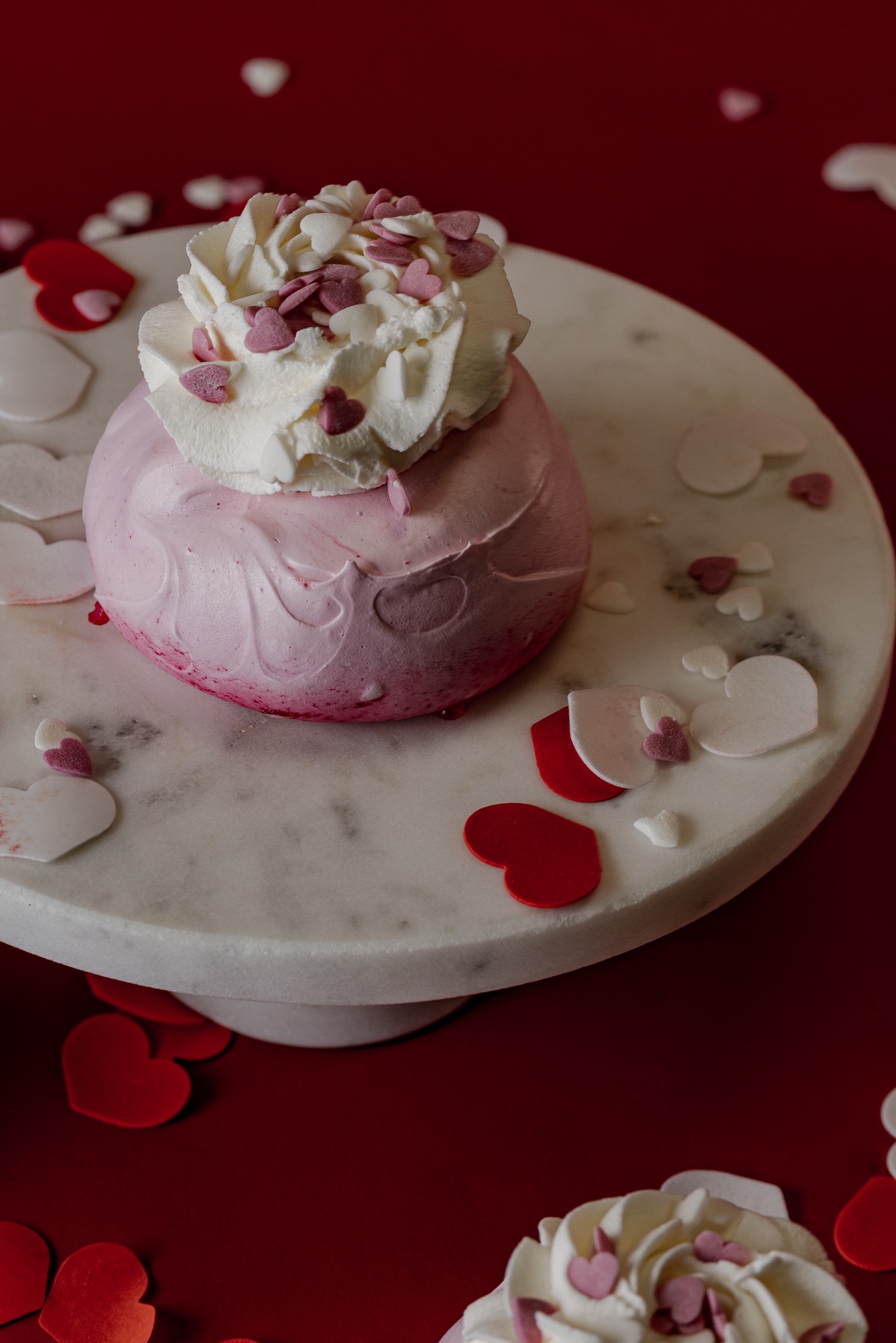Różowy deser bezowy z bitą śmietaną i dekoracjami w kształcie serc na marmurowej paterze, na czerwonym tle z serduszkami.