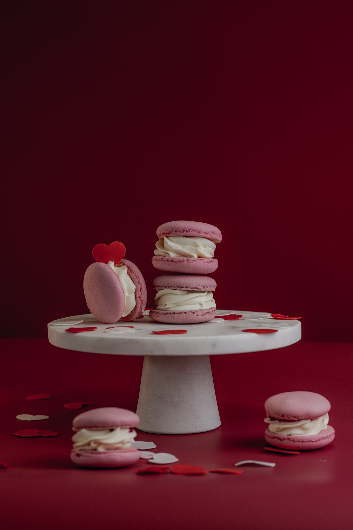 Różowe makaroniki z kremem na marmurowym stojaku, jeden z dekoracją w kształcie czerwonego serca, otoczone konfetti w kształcie serc na tle bordowym.