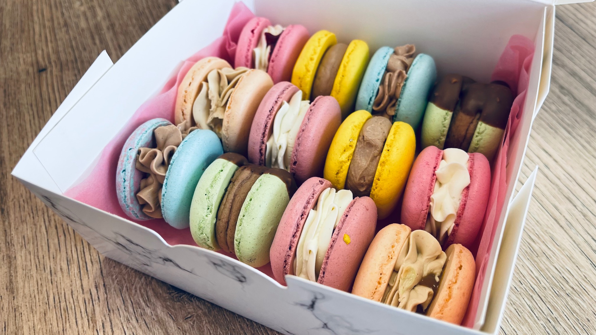 Różnokolorowe makaroniki z kremowymi nadzieniami w białym pudełku wyłożonym różowym papierem, widoczne na drewnianym blacie.