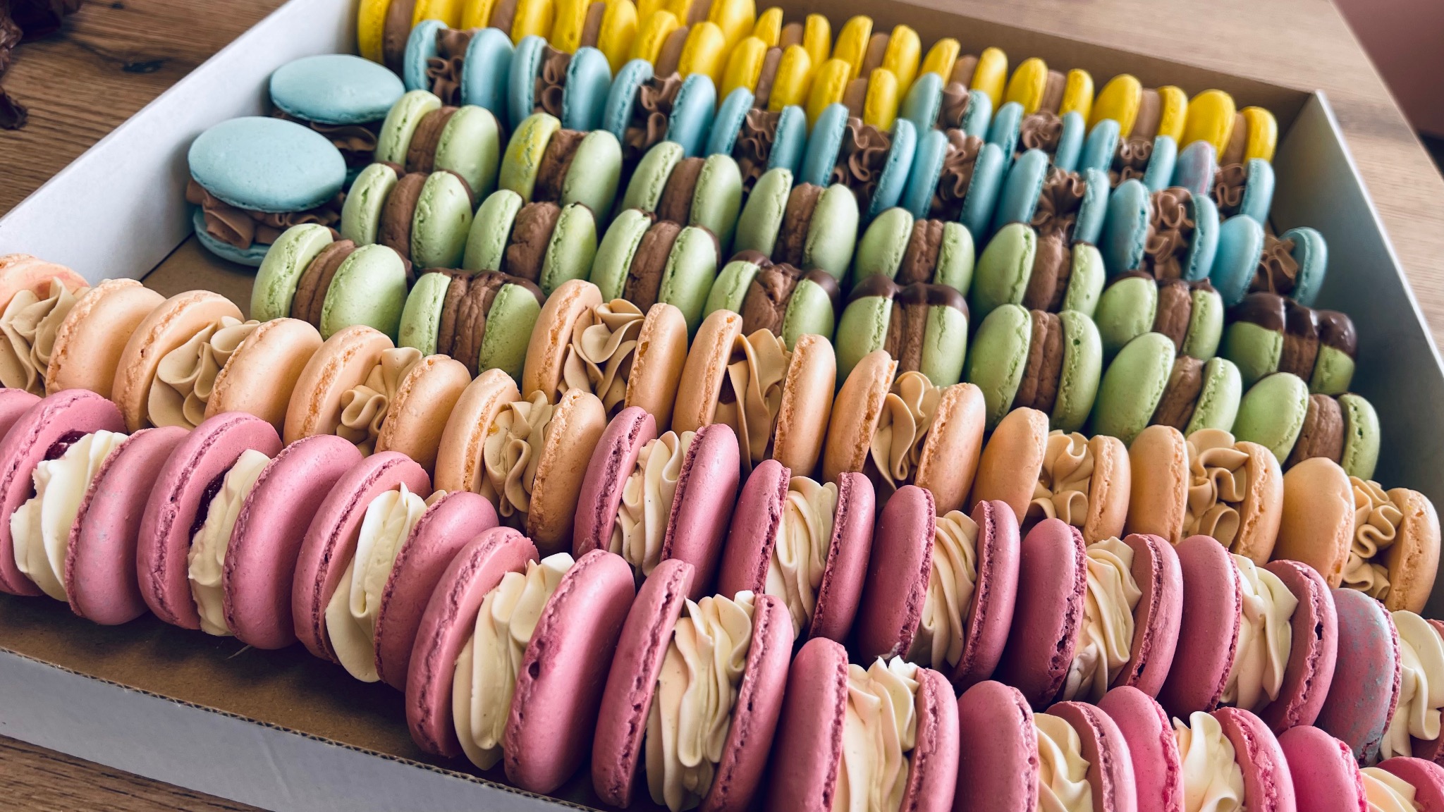 Rządki kolorowych makaroników z kremowymi nadzieniami w kartonowym pudełku, widoczne odcienie różu, beżu, zieleni, błękitu i żółci.