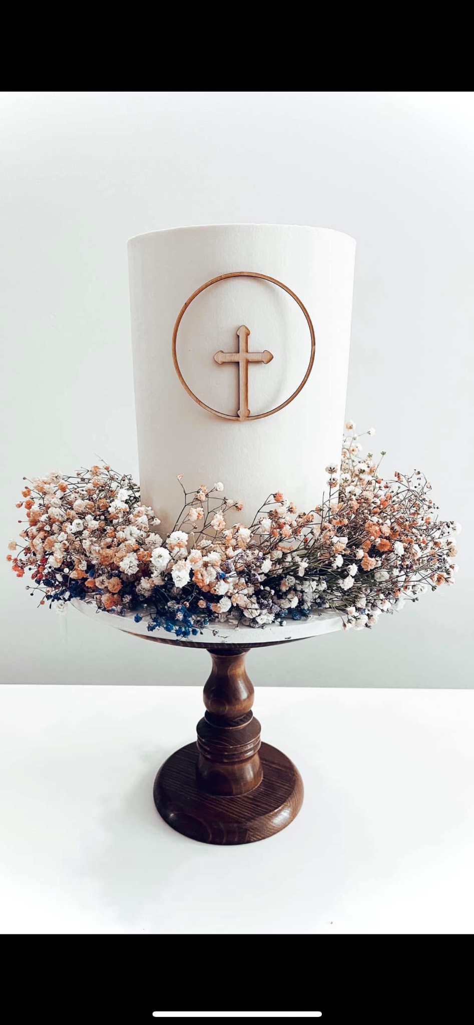 Tort komunijny z drewnianym krzyżem w okręgu, otoczony wiankiem z gipsówki w odcieniach bieli, pomarańczy i błękitu, ustawiony na drewnianej paterze na białym tle.