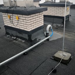 Andrzej Nowacki - Widok z góry na płaski dach z instalacjami odgromowymi, wentylacyjnymi i okablowaniem w korytkach. Na ceglanych kominach zamontowane metalowe obudowy wentylacyjne z żółtymi skrzynkami elektrycznymi...