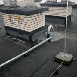 Widok z góry na płaski dach z instalacjami odgromowymi, wentylacyjnymi i okablowaniem w korytkach. Na ceglanych kominach zamontowane metalowe obudowy wentylacyjne z żółtymi skrzynkami elektrycznymi...