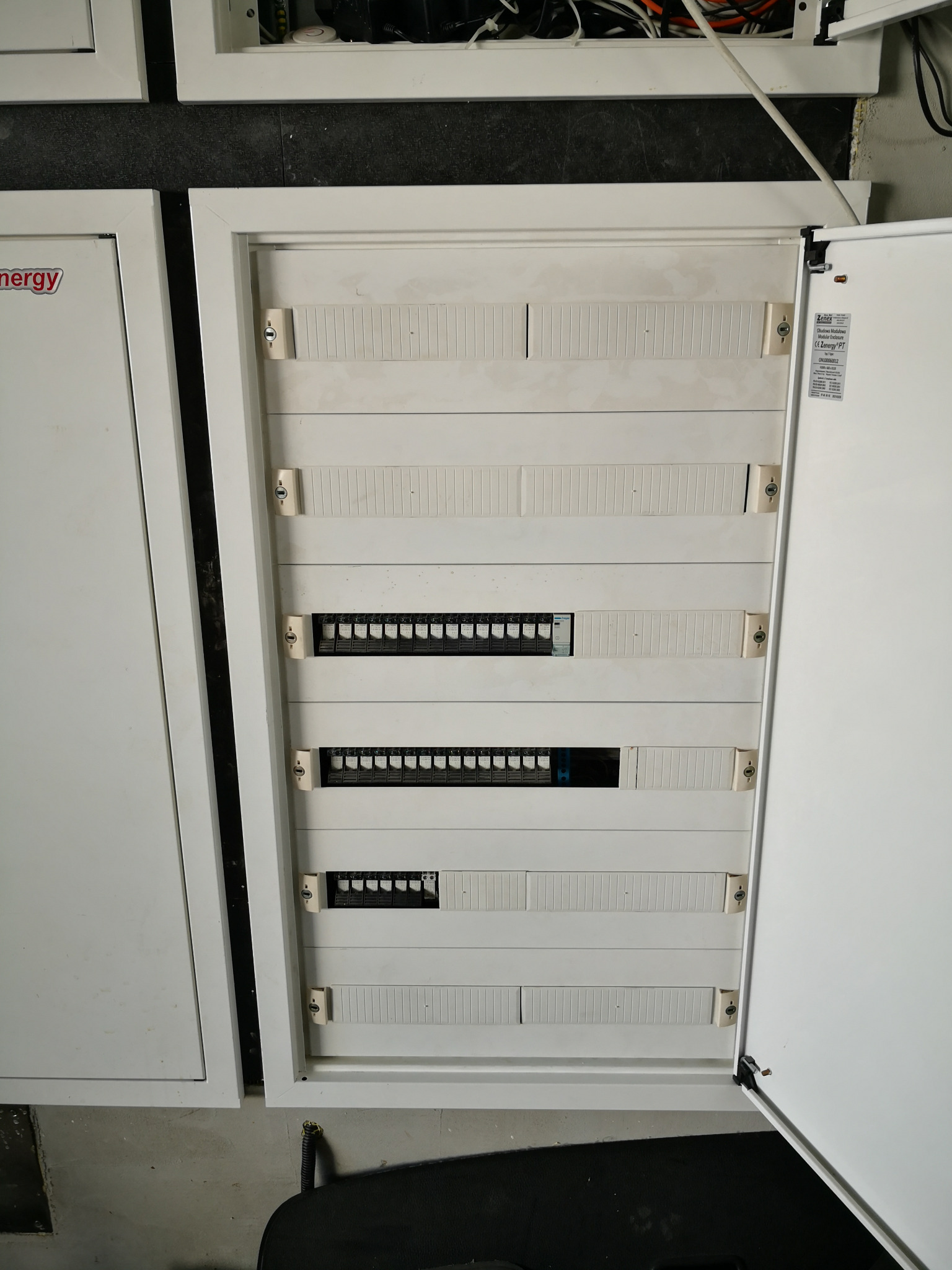 Otwarta szafa sterownicza Zenergy z modułami INT-O i INT-E oraz stycznikami, przeznaczona do sterowania oświetleniem w domu, widok z góry na szyny DIN z zamontowanymi zabezpieczeniami.