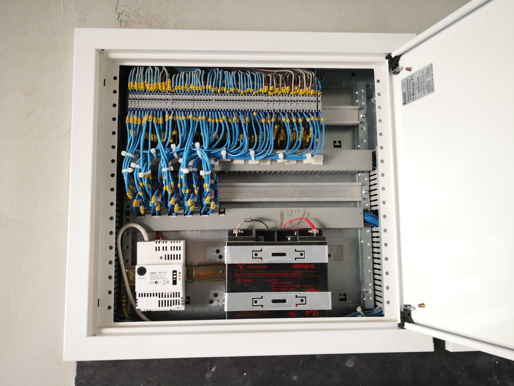 Otwarta szafa sterownicza z szynami WAGO, niebieskimi przewodami, modułem buforowym SATEL APS-412 i akumulatorem HPB18-12, widoczne złącza i oznaczenia.