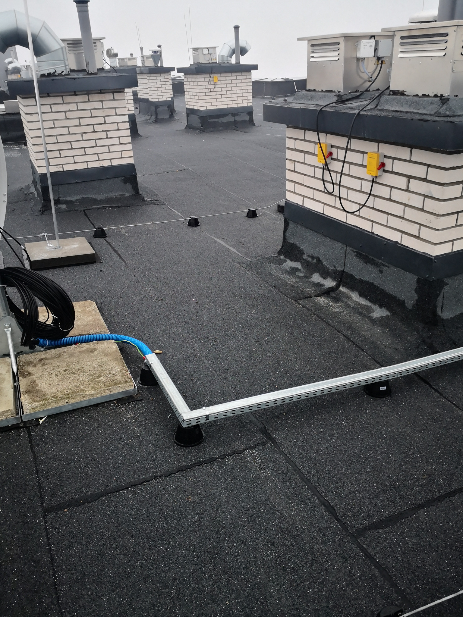 Instalacja odgromowa na dachu budynku z ceglanymi kominami wentylacyjnymi, widoczne przewody i uchwyty mocujące.