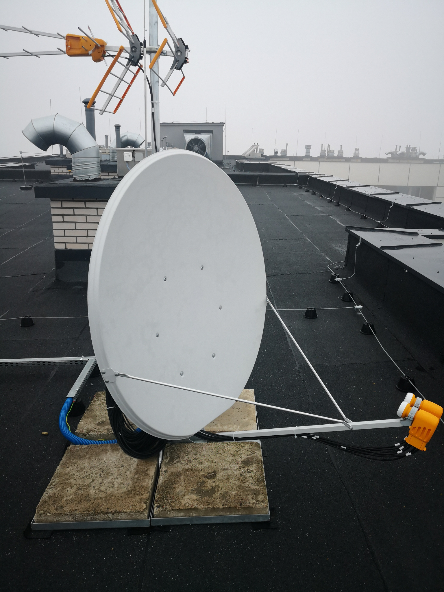 Antena satelitarna zamontowana na płaskim dachu, widoczne kable i elementy mocujące, w tle inne anteny i kominy wentylacyjne.