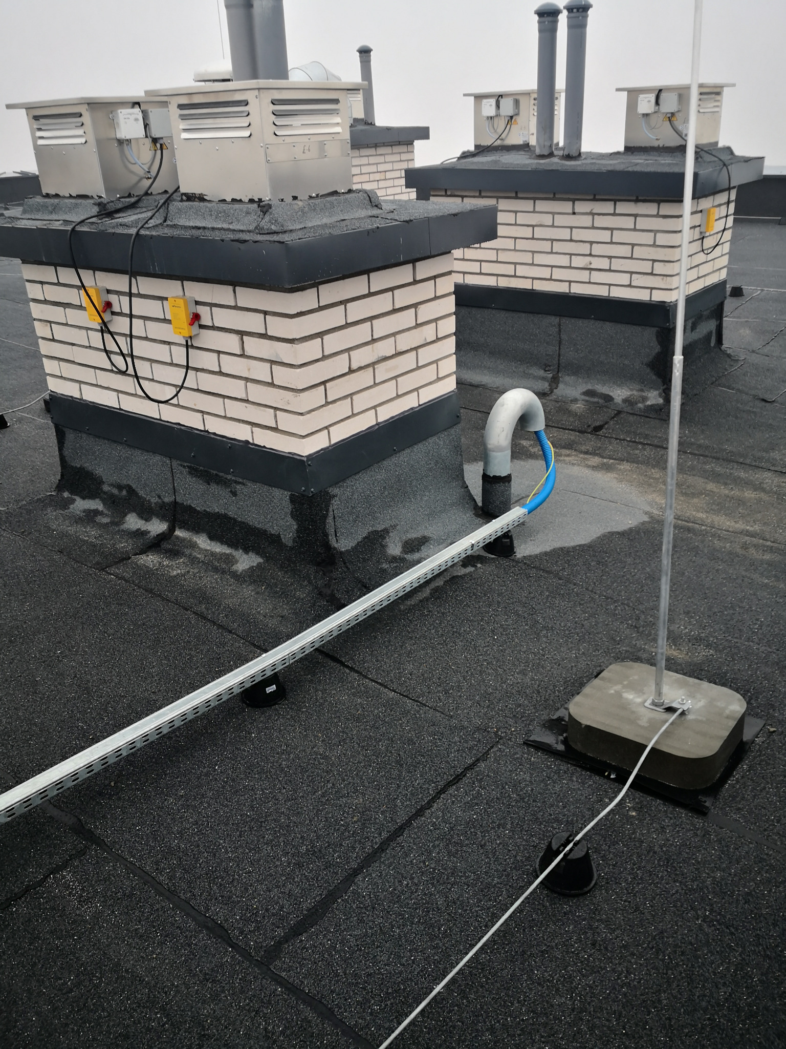 Widok z góry na płaski dach z instalacjami odgromowymi, wentylacyjnymi i okablowaniem w korytkach. Na ceglanych kominach zamontowane metalowe obudowy wentylacyjne z żółtymi skrzynkami elektrycznymi...