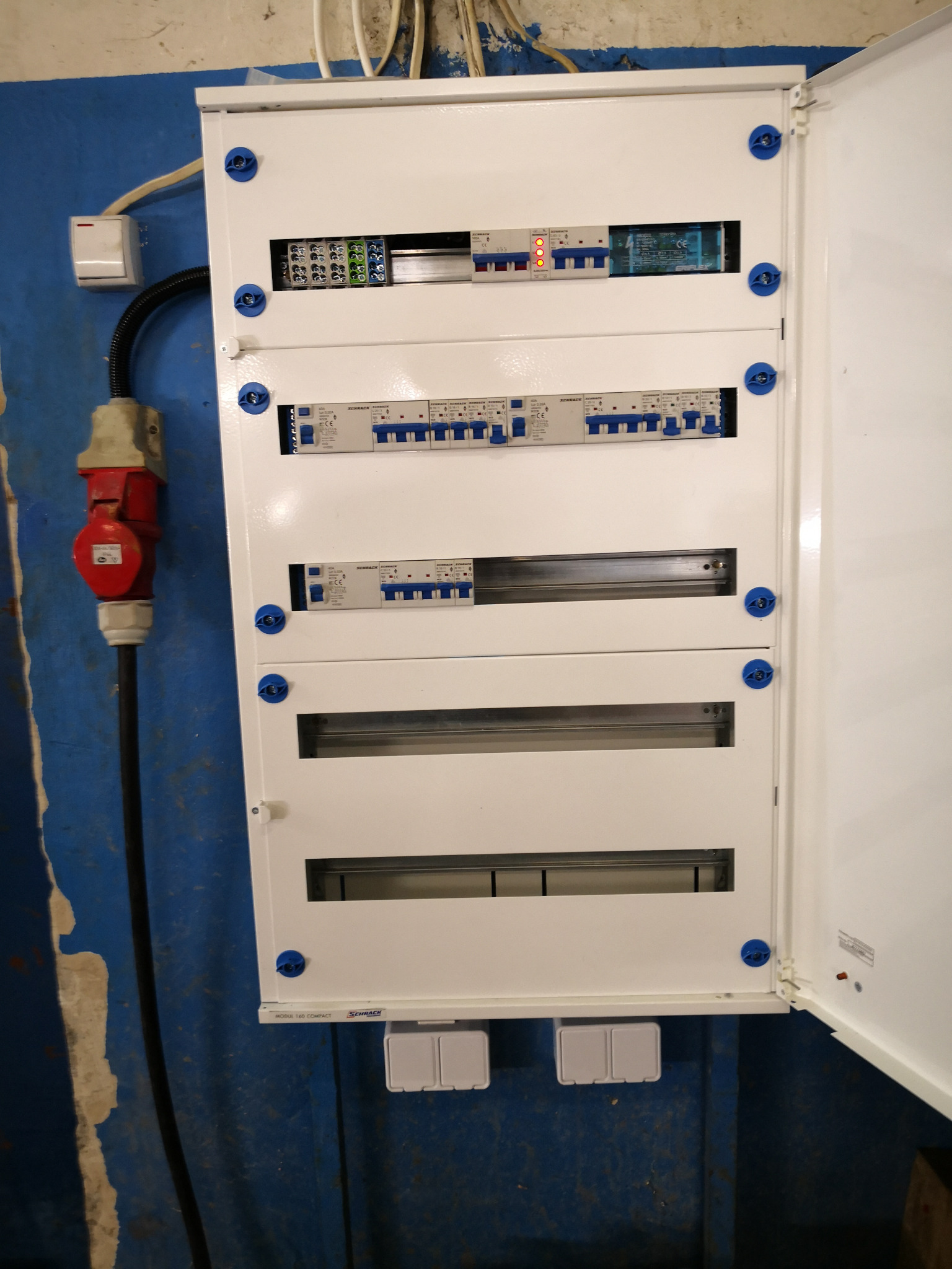 Nowoczesna, biała rozdzielnia elektryczna z widocznymi bezpiecznikami i elementami instalacji, zamontowana na niebieskiej, surowej ścianie w hali przemysłowej. Widoczny czerwony wtyk siłowy po...