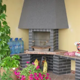 Murowany grill z szarej cegły z blatem roboczym, otoczony roślinami w donicach i zapasem wody, na brązowej terakocie.