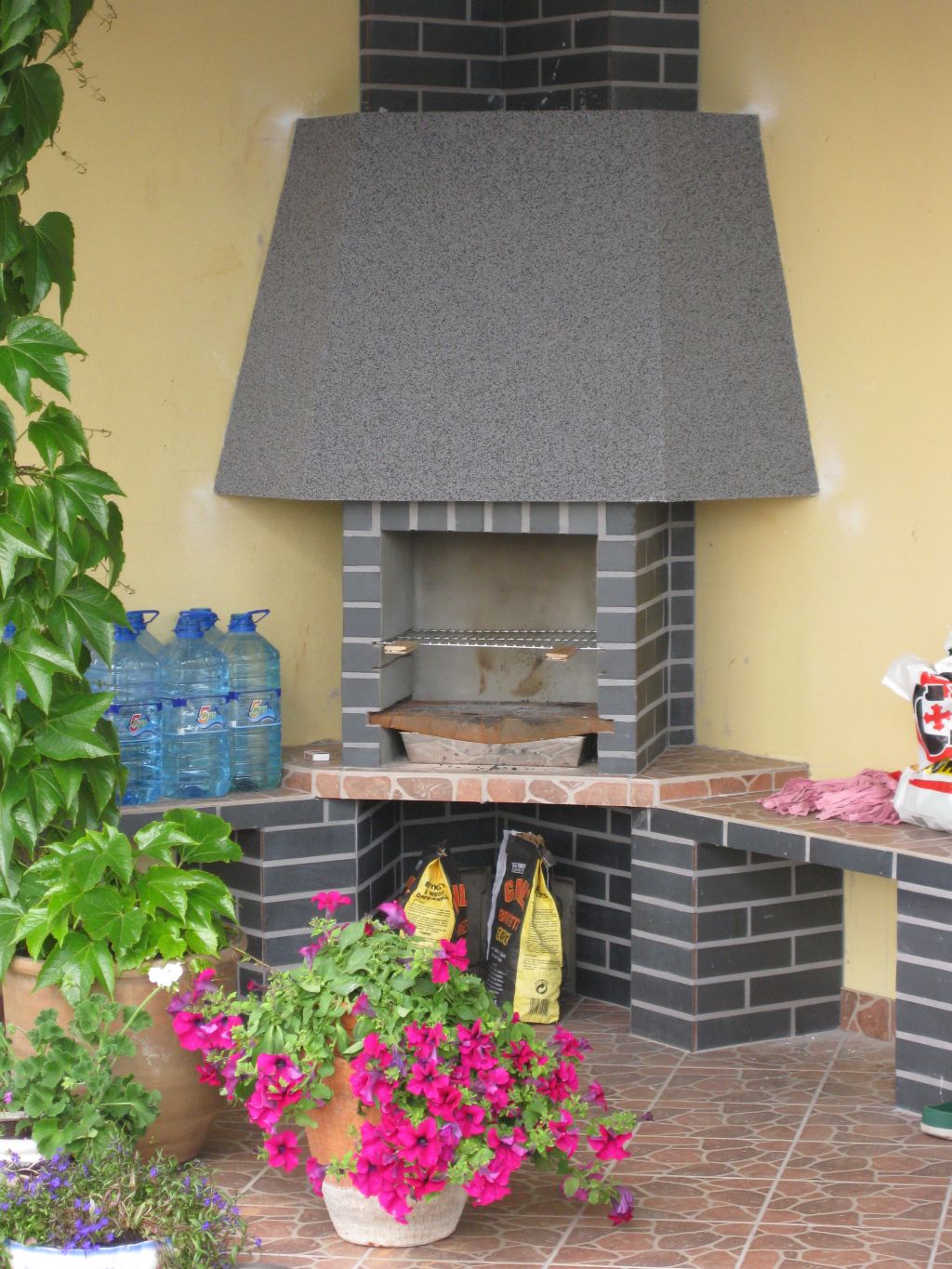 Murowany grill z szarej cegły z blatem roboczym, otoczony roślinami w donicach i zapasem wody, na brązowej terakocie.