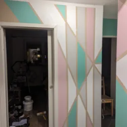 Ściana pomalowana w geometryczne wzory w pastelowych kolorach: różowy, miętowy i beżowy, widoczne narzędzia malarskie i bałagan w tle.