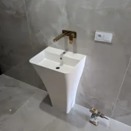 Biała, nowoczesna umywalka kolumnowa z baterią w kolorze złotym, zamontowana na tle szarych, marmurkowych płytek. Obok umywalki leży wkrętarka.