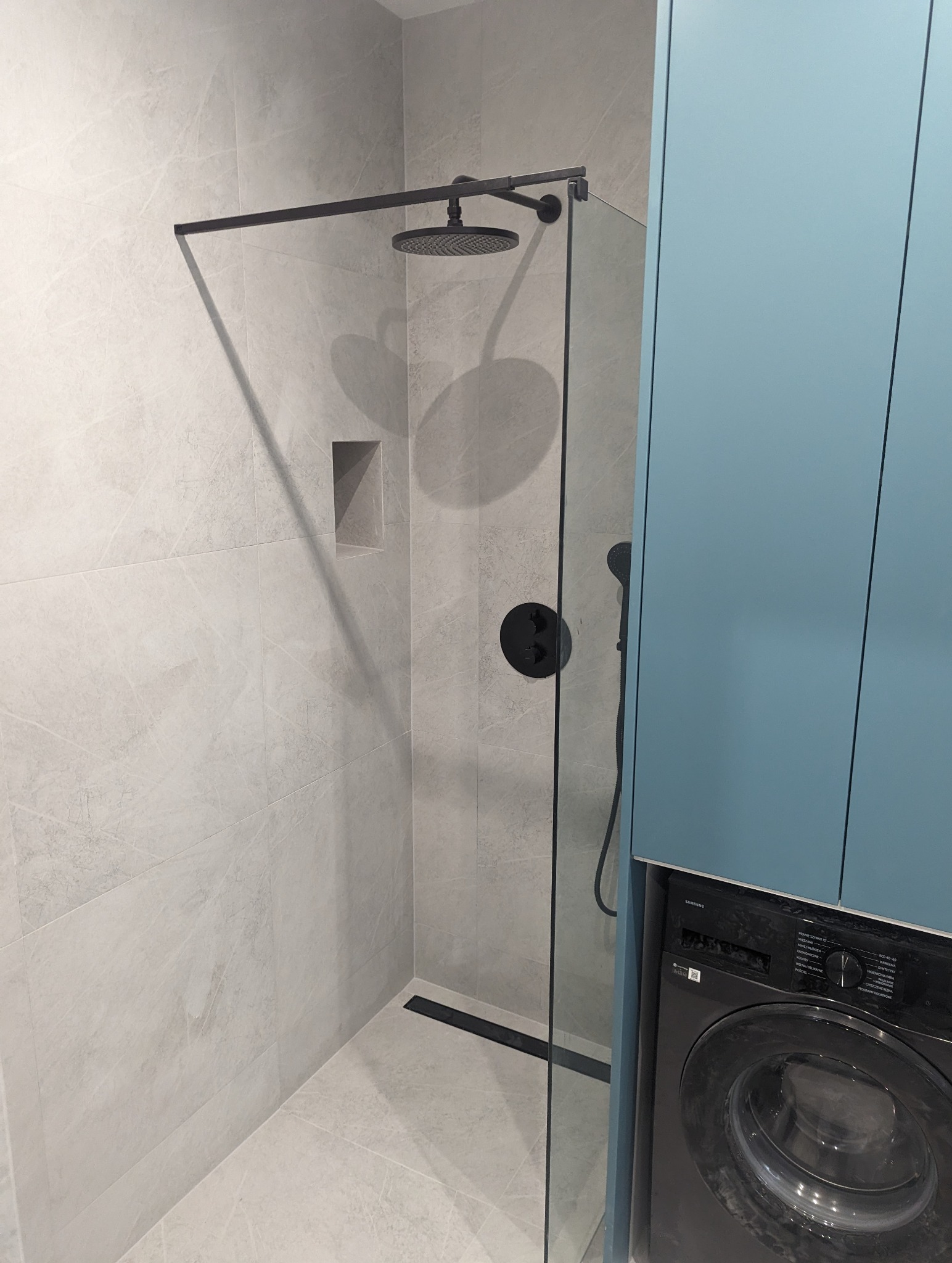 Nowoczesna kabina prysznicowa z czarną armaturą, odpływem liniowym i szarymi płytkami imitującymi marmur, obok pralka zabudowana w szafce w kolorze błękitnym.
