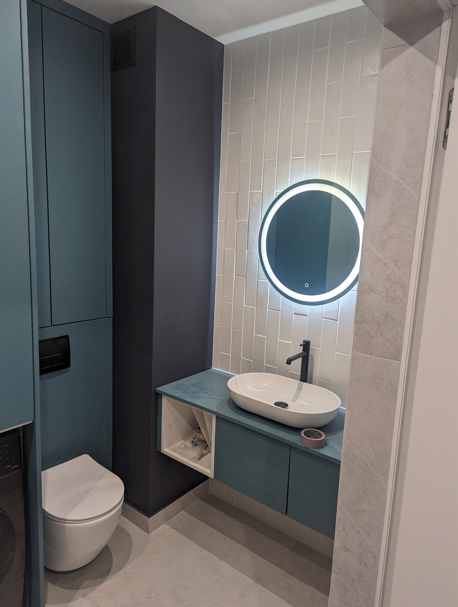 Nowoczesna łazienka z podświetlanym lustrem, wiszącą szafką z umywalką, toaletą i białymi płytkami ułożonymi pionowo, widoczne narzędzia w szafce.