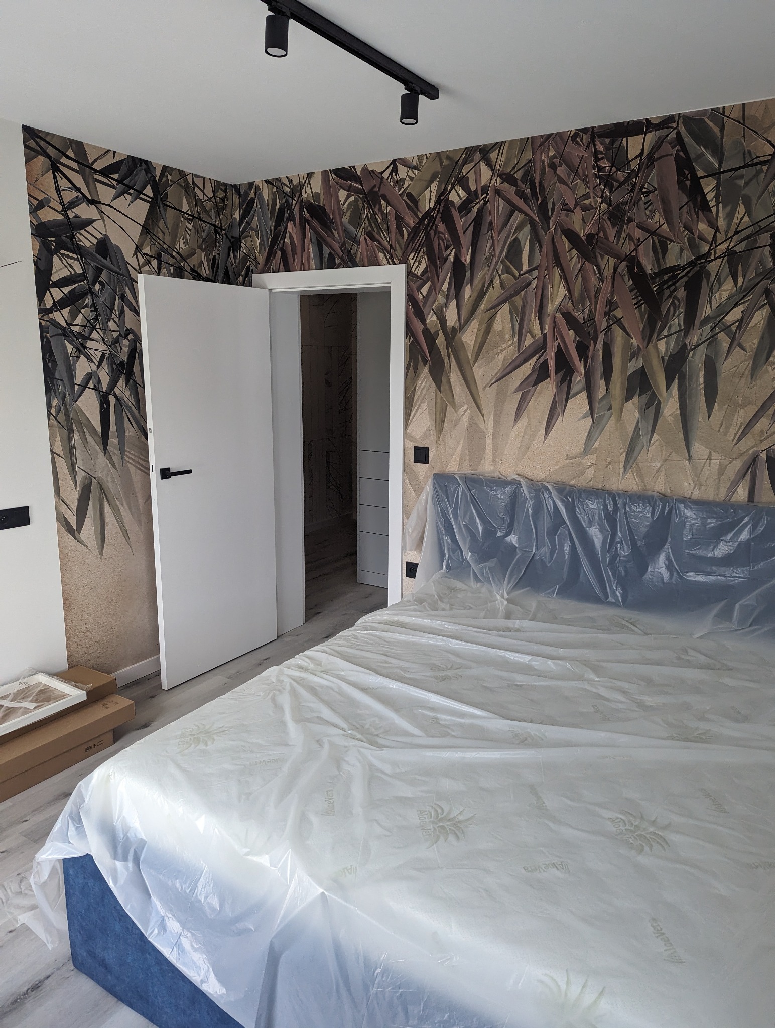Pokój z tapetą w liście bambusa i świeżo zamontowanymi drzwiami, łóżko przykryte folią ochronną, widoczne kartony po panelach podłogowych.