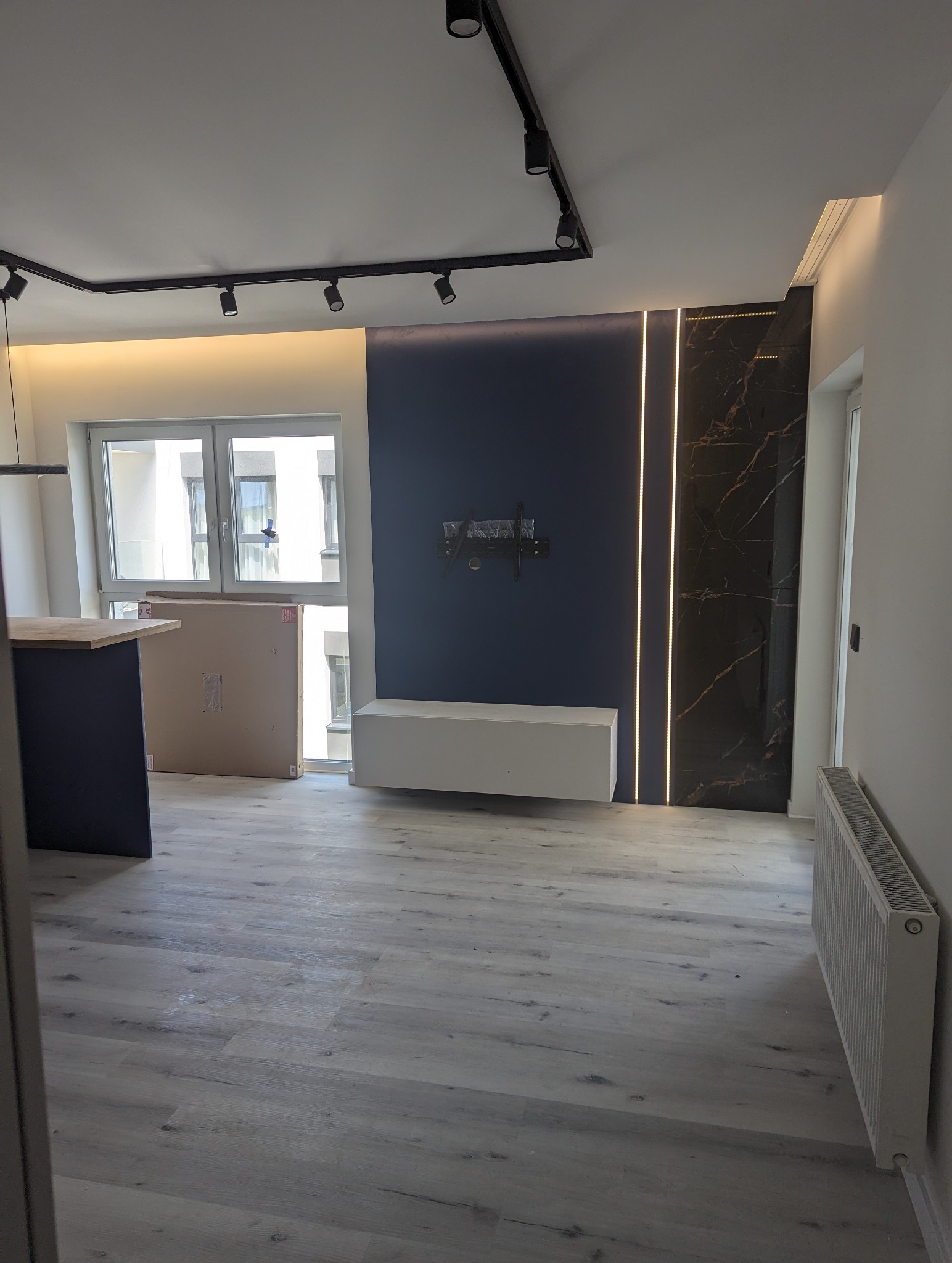 Nowoczesne wnętrze w trakcie aranżacji: szare panele podłogowe, ciemnoniebieska ściana z podświetleniem LED, biała szafka RTV i zamontowany uchwyt na telewizor, widoczne kartony z wyposażeniem...