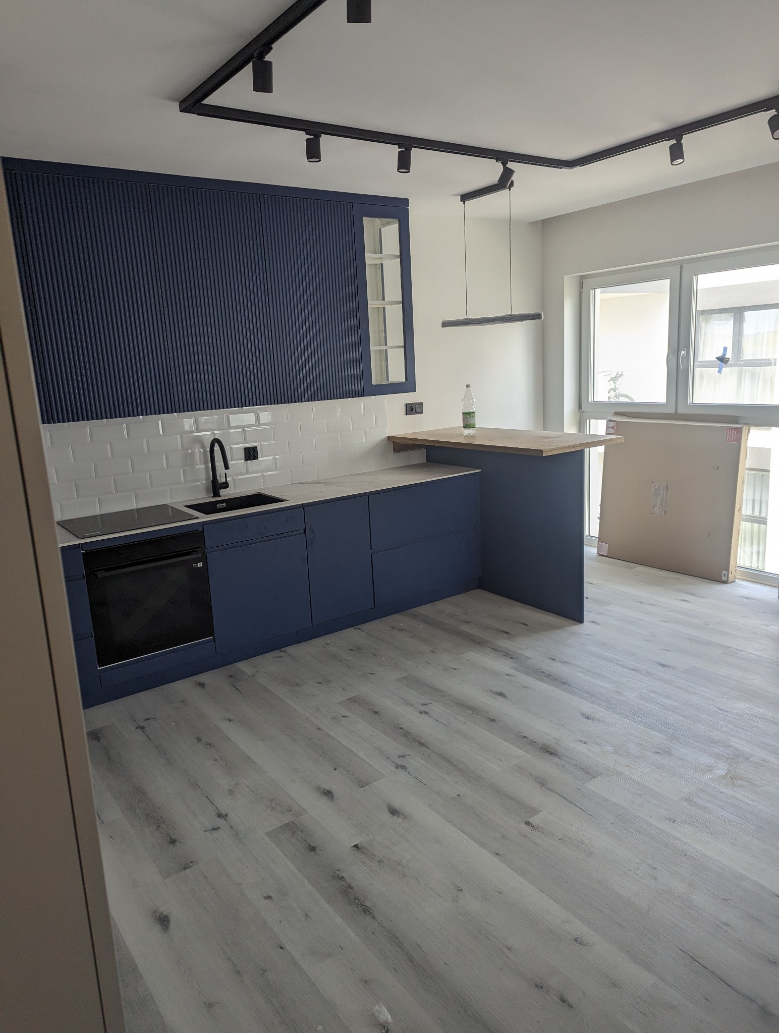 Nowoczesna kuchnia w odcieniach granatu i bieli, z ryflowanymi frontami szafek, białymi płytkami metra i drewnianym blatem baru, oświetlona reflektorami szynowymi.