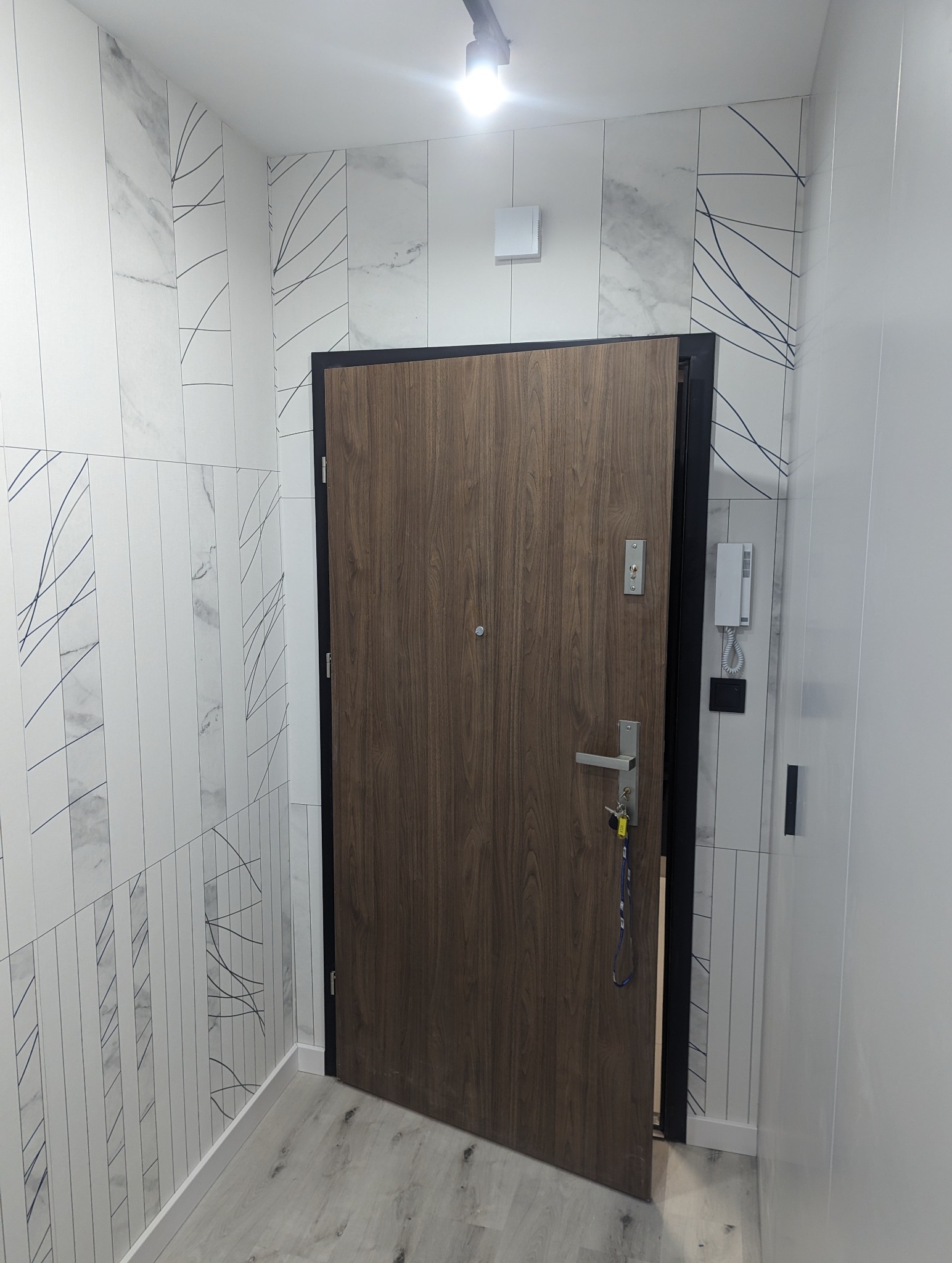 Nowoczesne wejście do mieszkania: drewniane drzwi z czarną ramą, jasne płytki ścienne z marmurowym wzorem i delikatnymi czarnymi liniami, szara podłoga, biały domofon