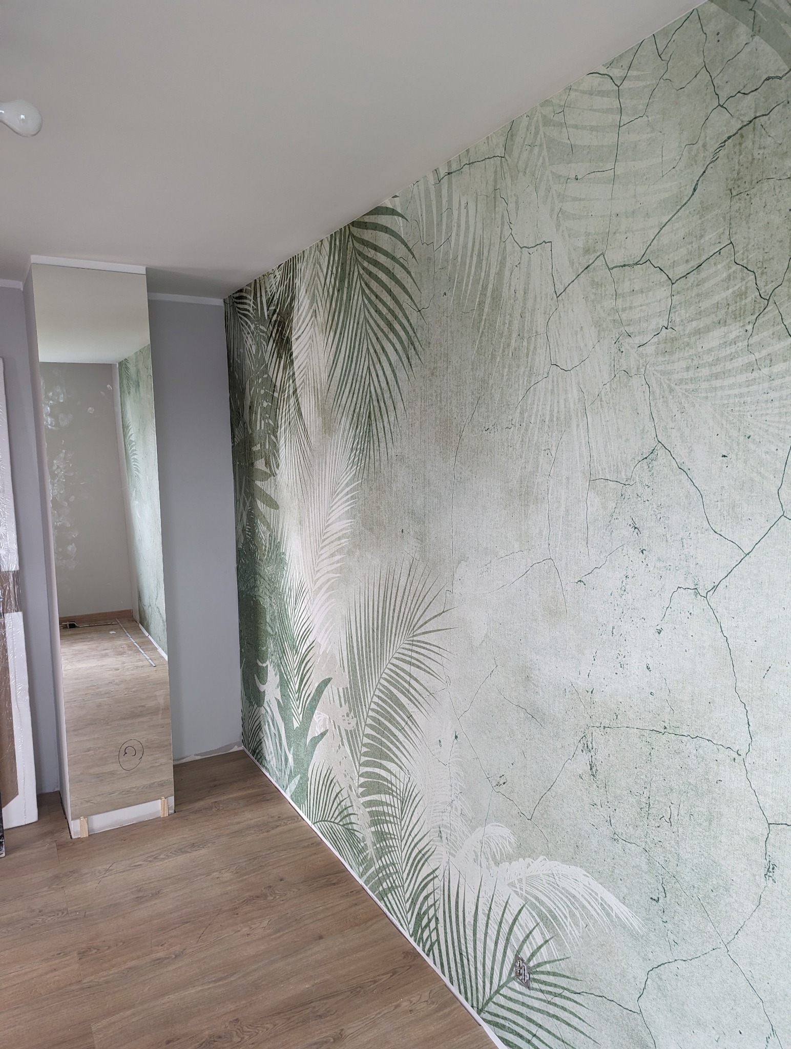 Ściana z tapetą w motywy roślinne, imitująca spękaną powierzchnię, widok na fragment podłogi z paneli i przejście do innego pomieszczenia w trakcie prac.