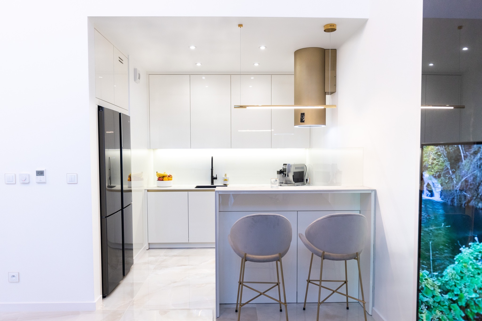 Nowoczesna, jasna kuchnia z białymi lakierowanymi frontami, wyspą z dwoma hokerami na złotych nogach, czarną lodówką i złotym okapem.