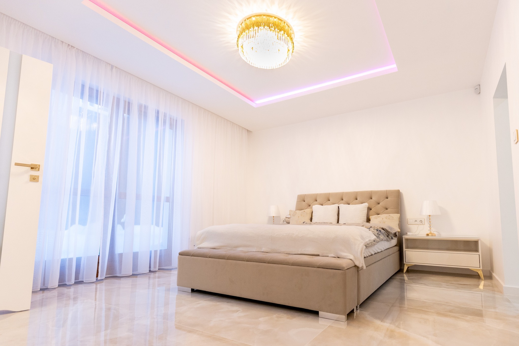 Jasna sypialnia z beżowym łóżkiem z pikowanym zagłówkiem, marmurową podłogą, kryształowym żyrandolem i podświetleniem LED na suficie.