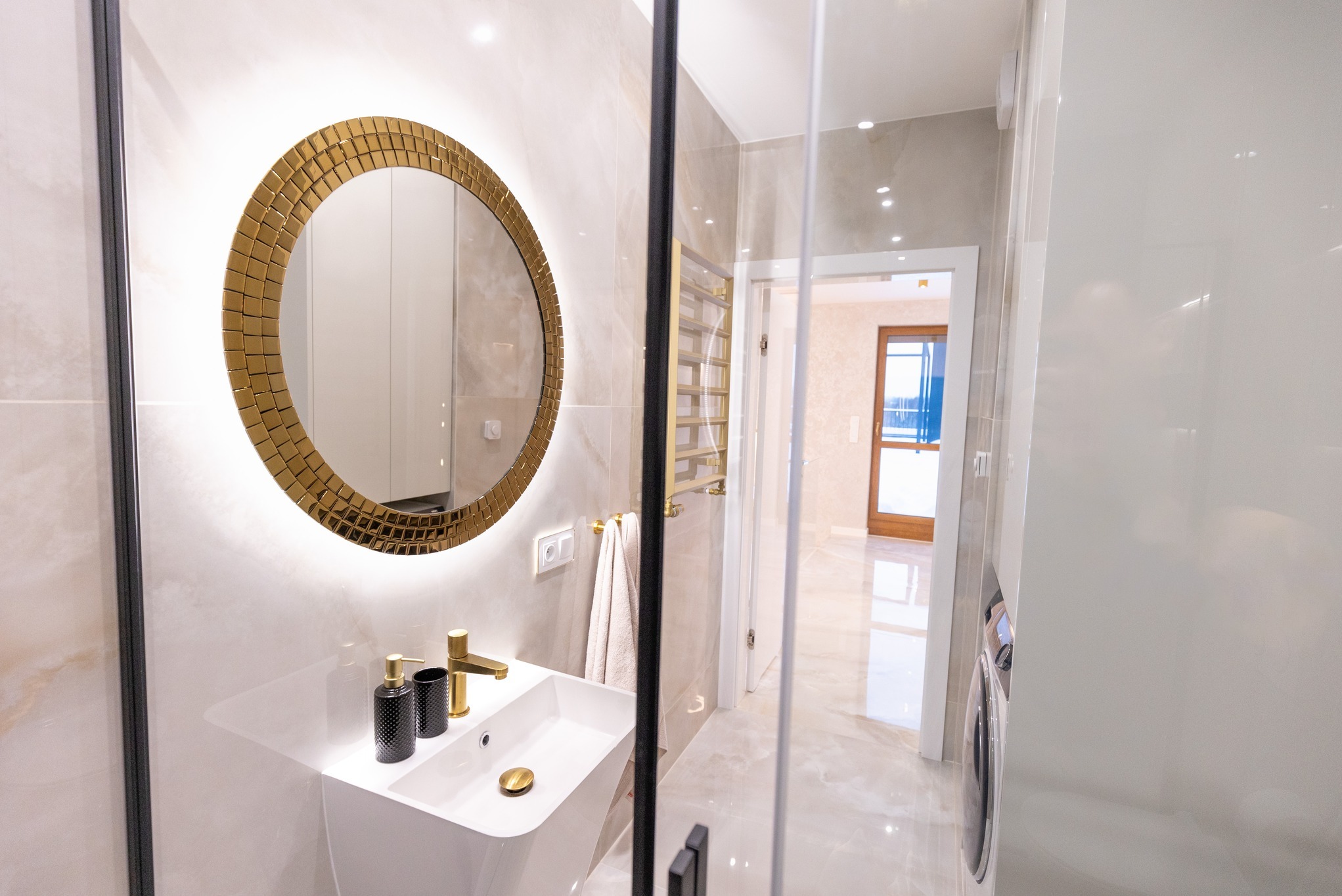 Nowoczesna łazienka z okrągłym lustrem podświetlanym LED, umywalką nablatową, złotą armaturą i czarnymi akcesoriami, widok przez szklaną kabinę prysznicową na dalszą część wykończonego wnętrza.