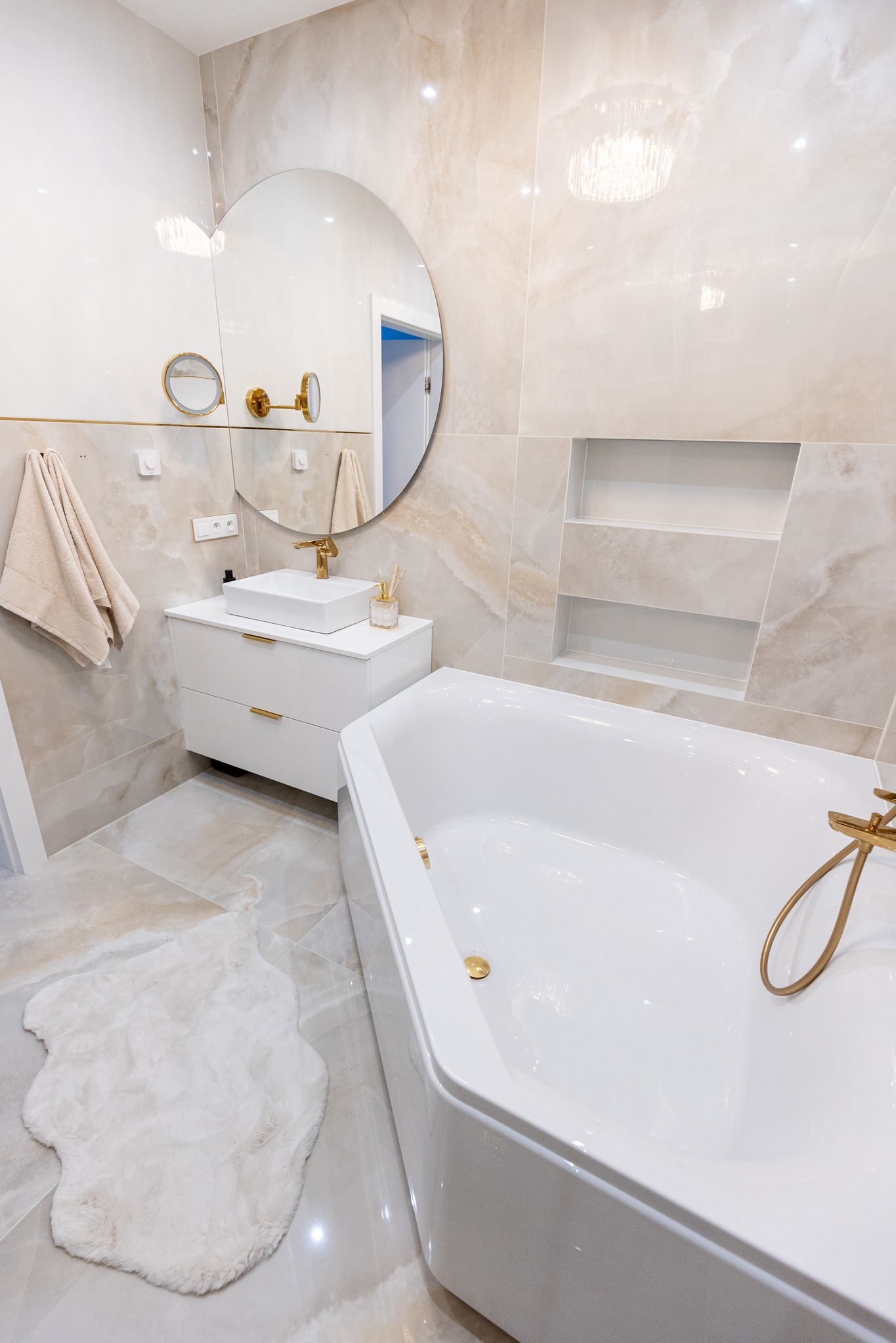 Jasna łazienka z beżowymi płytkami imitującymi marmur, białą wanną narożną, szafką z umywalką i okrągłym lustrem z pozłacanymi detalami.