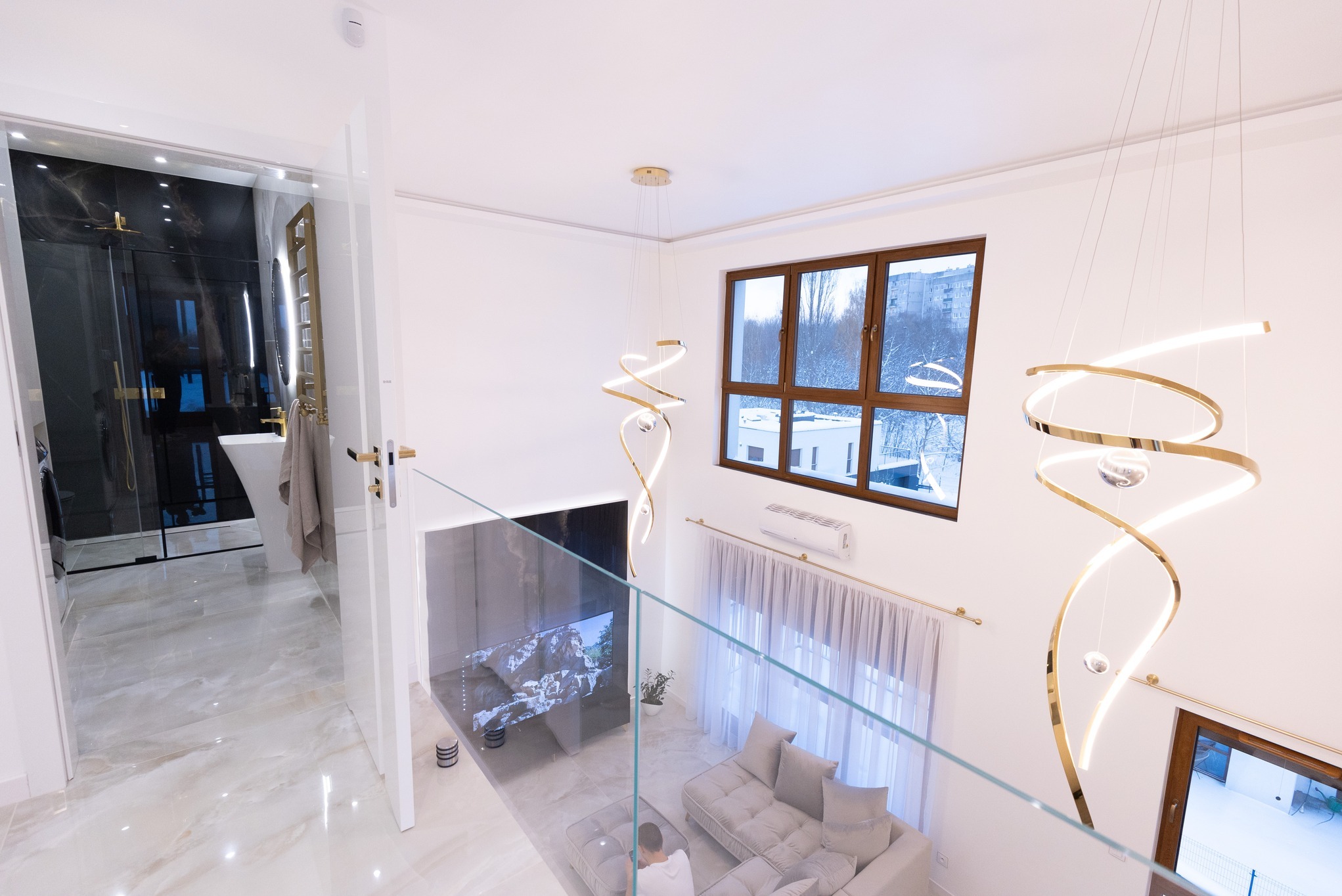 Dwupoziomowe wnętrze domu z widokiem na łazienkę z czarnymi płytkami, szklaną kabiną prysznicową i wolnostojącą umywalką, marmurowe płytki podłogowe, salon z szarą sofą i telewizorem, nowoczesne...