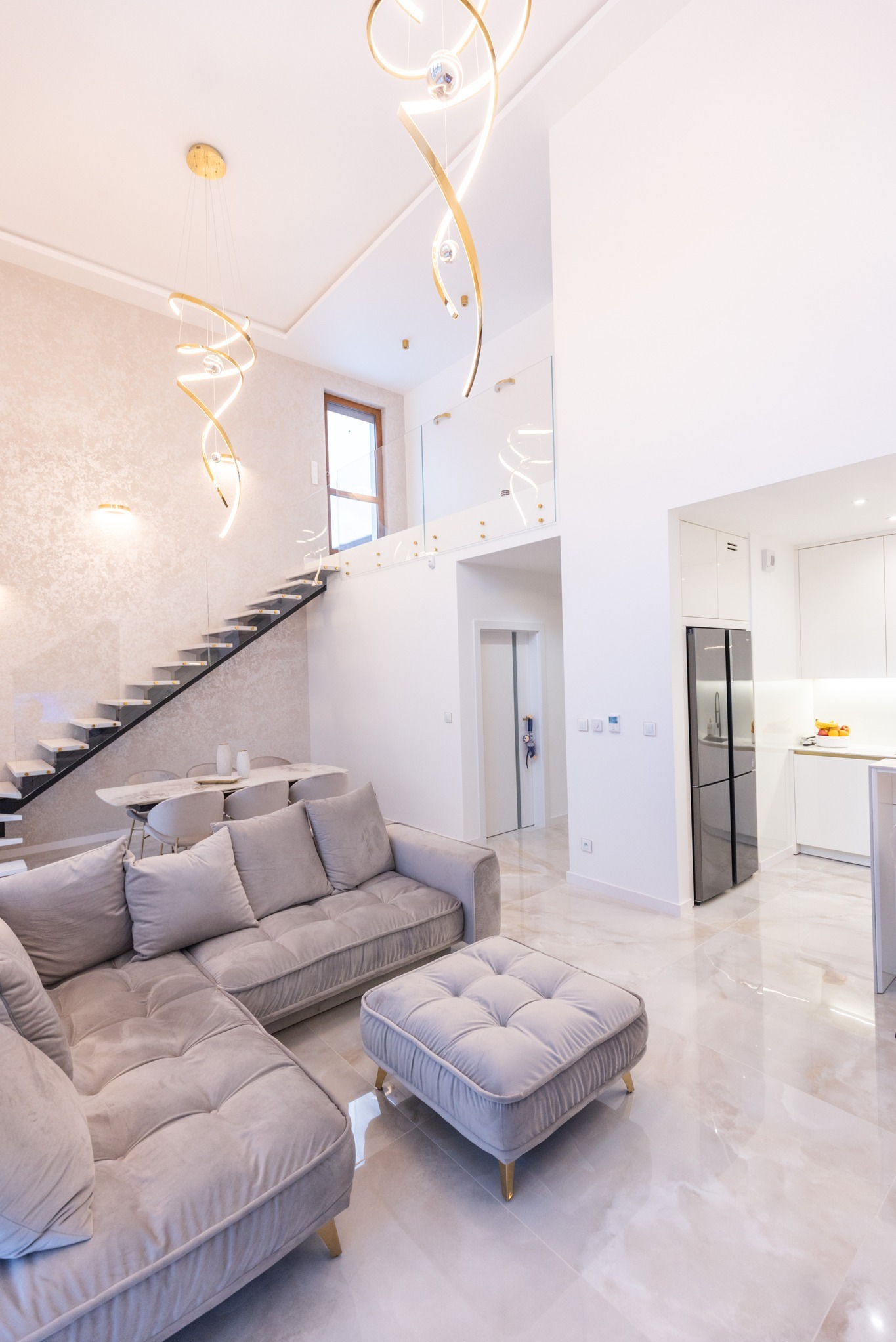Salon z antresolą i schodami, szara sofa narożna z pufą, marmurowe płytki podłogowe, nowoczesne, złote lampy wiszące, biała kuchnia w tle, jasne wnętrze.
