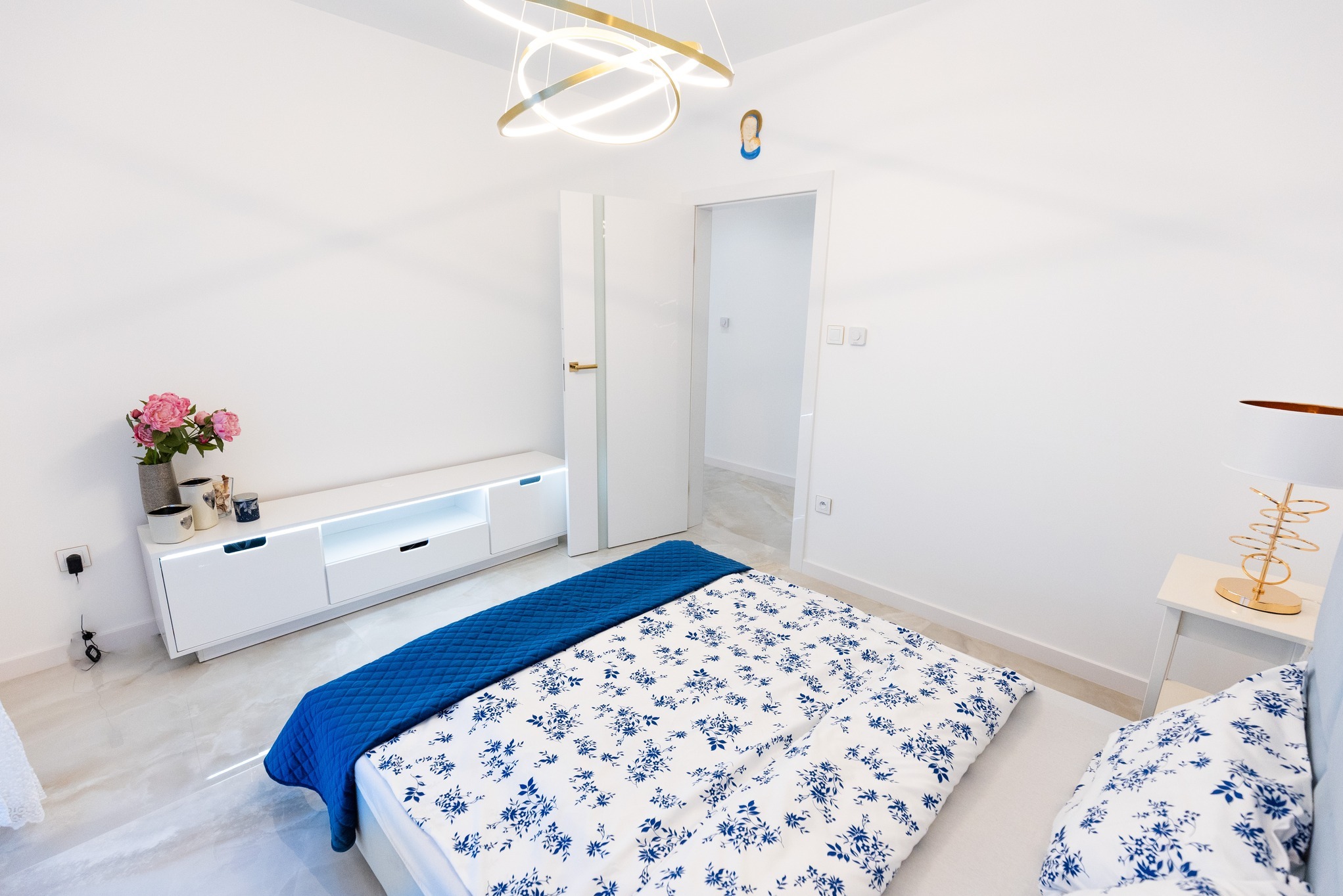 Nowoczesna sypialnia z białymi ścianami, marmurową podłogą, łóżkiem z niebieską narzutą w kwiaty, białą szafką RTV z podświetleniem LED, kwiatami w wazonie i minimalistyczną lampą wiszącą.