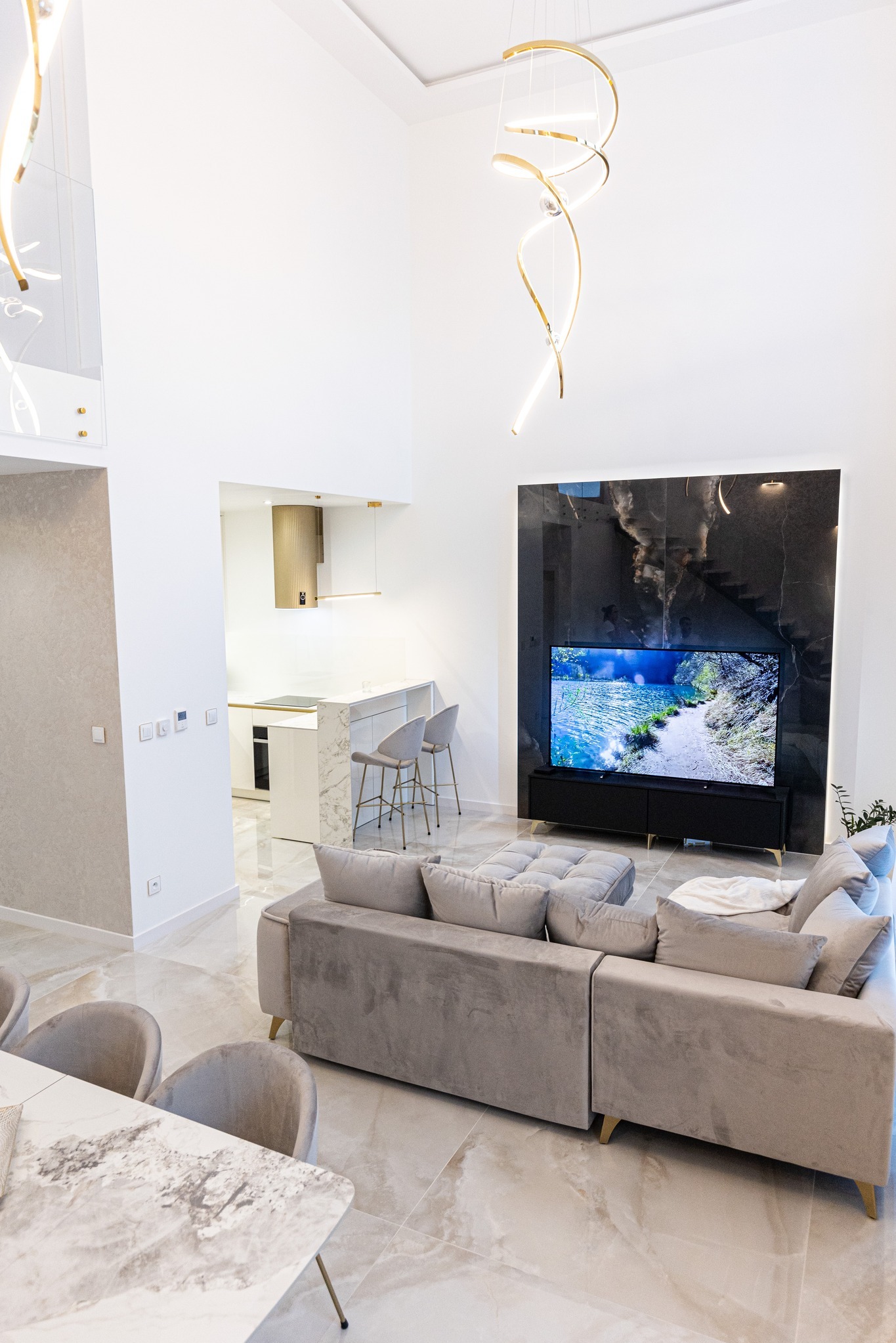 Jasne, przestronne wnętrze z marmurową podłogą i ścianą telewizyjną z czarnego marmuru, szary narożnik z poduszkami, otwarta kuchnia z marmurowym blatem i złotymi akcentami oraz nowoczesne, złote...