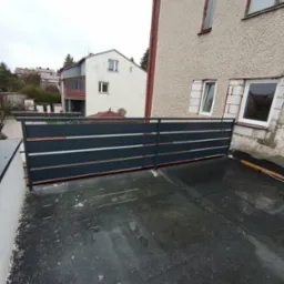 Szara, pozioma balustrada z metalowych profili zamontowana na płaskim dachu budynku wielorodzinnego, widok z góry na mokrą powierzchnię dachu.