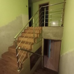 Jakub Wcisło - Drewniane schody wewnętrzne z balustradą ze stali nierdzewnej, widok z dołu, ściany w kolorze pistacjowym, widoczne niedokończone wykończenia ścian.