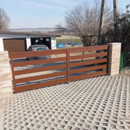 Nowoczesna, brązowa brama wjazdowa z poziomych profili, zamontowana na słupkach z imitacji kamienia. Widoczna kostka ażurowa na podjeździe, garaż z samochodem w tle i słupy energetyczne.