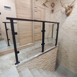 Jakub Wcisło - Szklana balustrada schodowa z czarnymi, metalowymi słupkami mocującymi, widok z góry na szare, kamienne schody wewnątrz domu, dekoracje myśliwskie na ścianie.