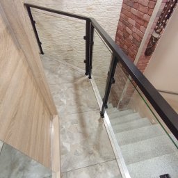 Jakub Wcisło - Widok z góry na schody z szarymi stopniami i szklaną balustradą z czarnymi elementami mocującymi, ściany wykończone w różnych stylach: cegła, kamień i drewno.