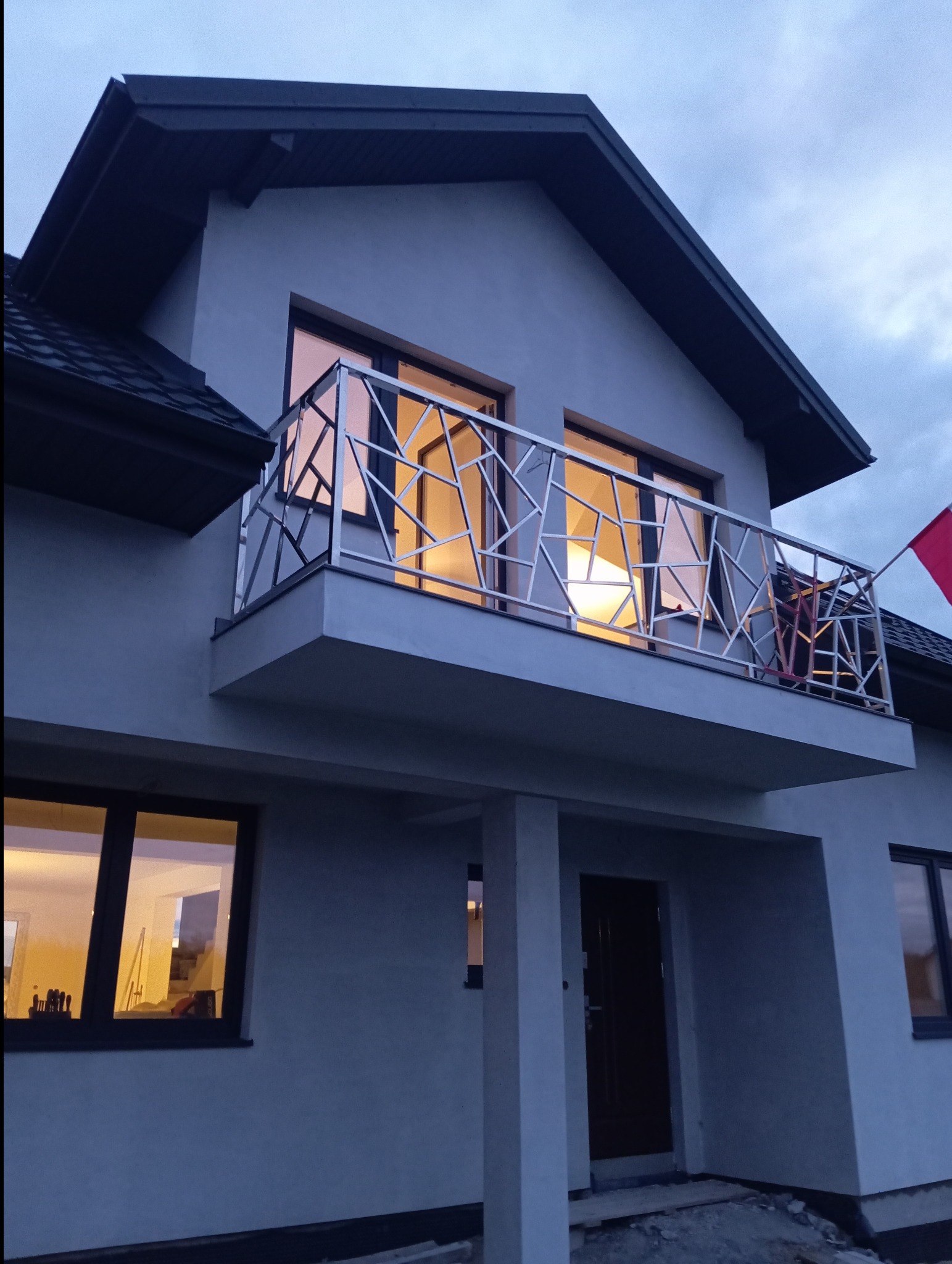 Nowoczesna balustrada balkonowa ze stali nierdzewnej o geometrycznym wzorze na tle elewacji domu z oknami, widoczna flaga Polski.