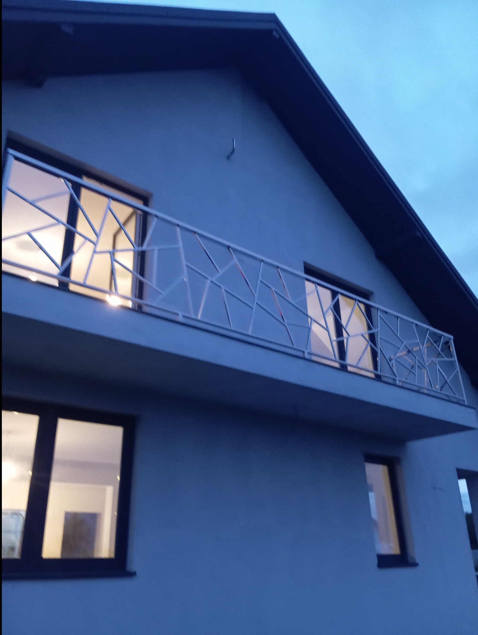 Nowoczesna balustrada balkonowa ze stali nierdzewnej o geometrycznym wzorze na tle szarej elewacji domu i wieczornego nieba.