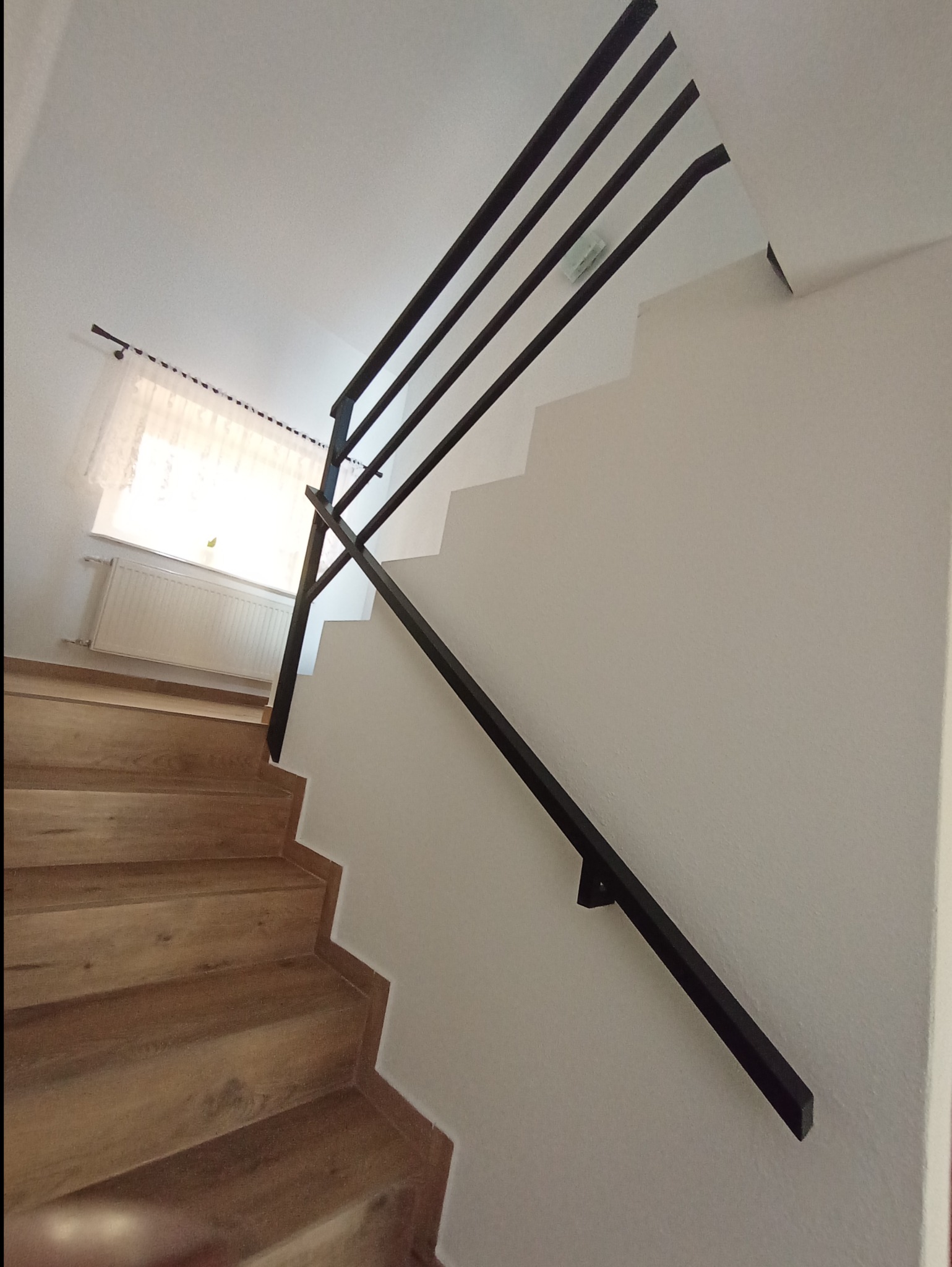 Wnętrze domu: Schody z drewnianymi stopniami i nowoczesną, minimalistyczną, czarną balustradą z metalowych prętów, zamontowaną do ściany.
