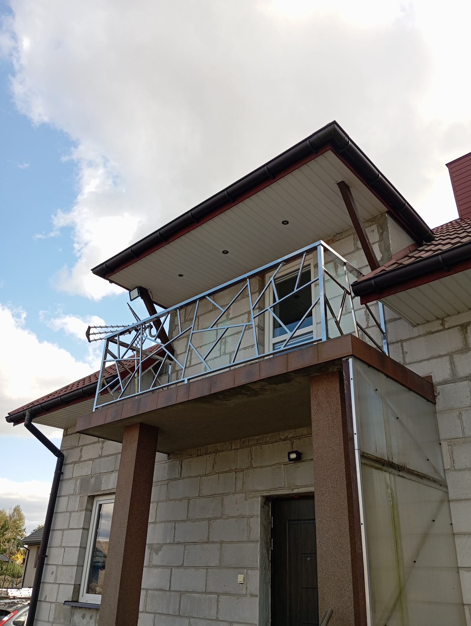 Nowoczesna balustrada balkonowa ze stali nierdzewnej o geometrycznym wzorze, zamontowana na balkonie domu z szarego bloczka betonowego, widok z dołu na tle błękitnego nieba z chmurami.