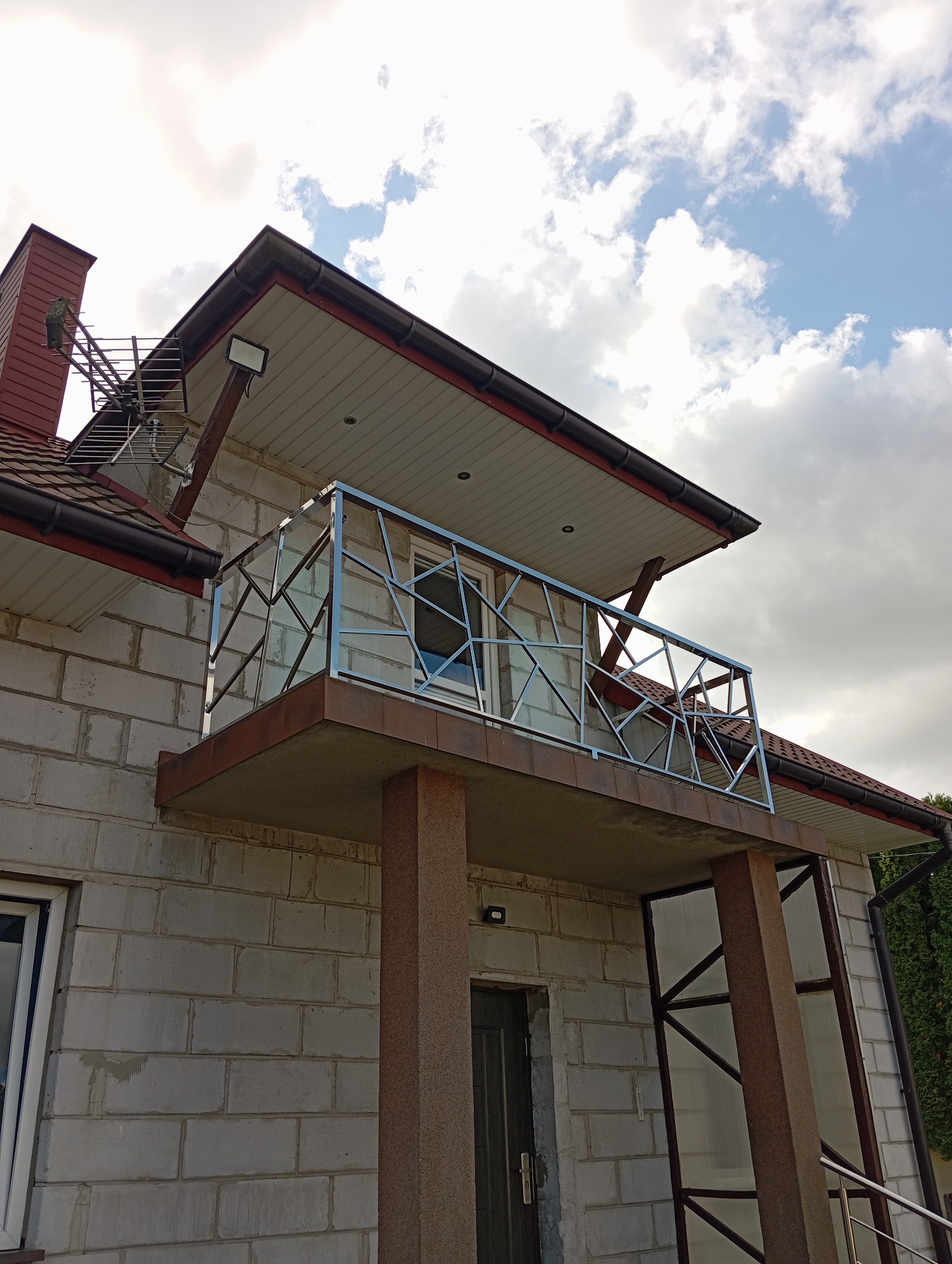 Balkon z nowoczesną balustradą nierdzewną o geometrycznym wzorze w Chęcinach, na tle surowej ściany z białego pustaka i błękitnego nieba z chmurami.