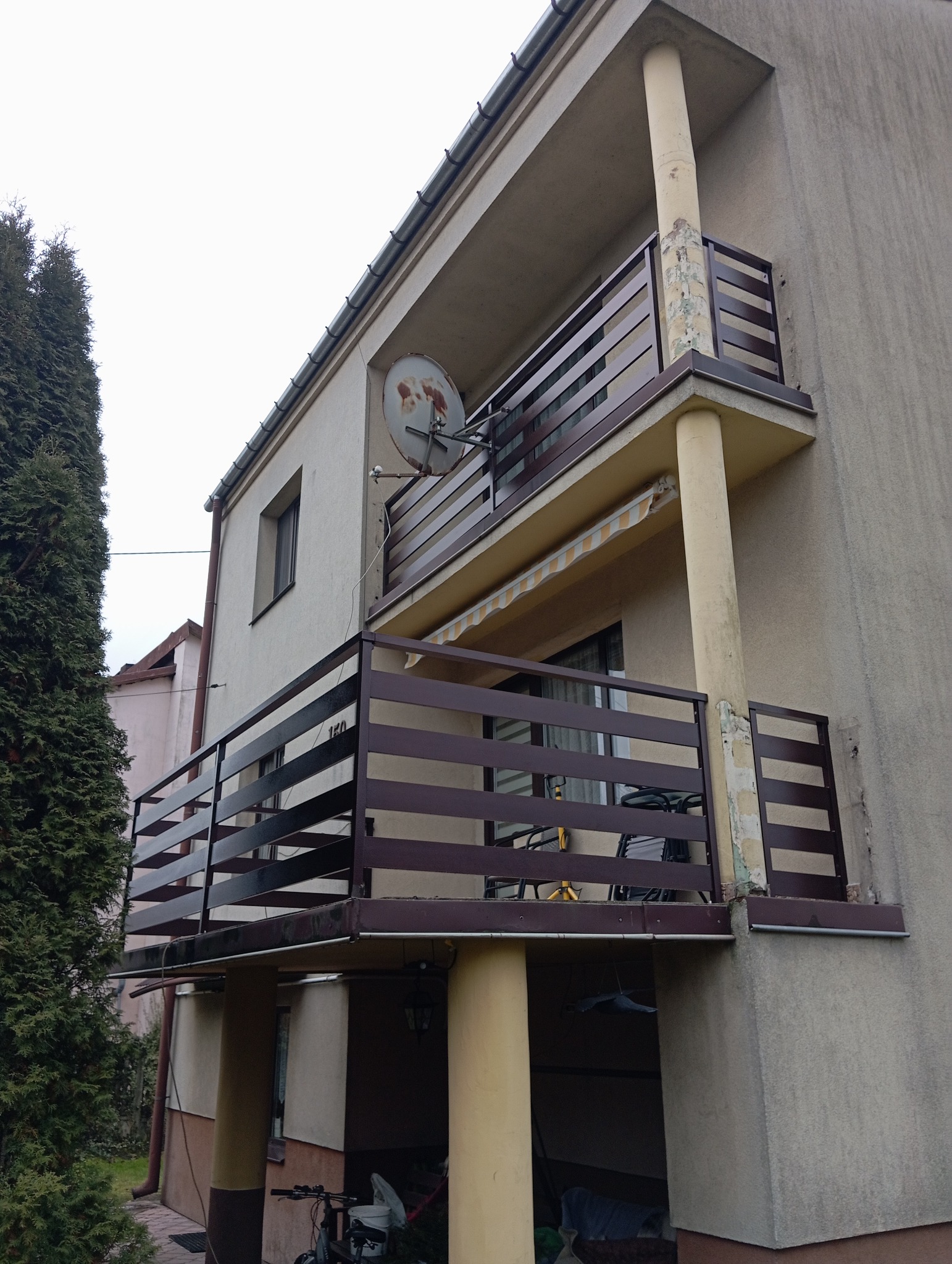 Dwukondygnacyjny budynek z balkonami zabezpieczonymi nowoczesnymi, poziomymi balustradami w kolorze ciemnego brązu. Na górnym balkonie widoczna antena satelitarna i zwinięta markiza.