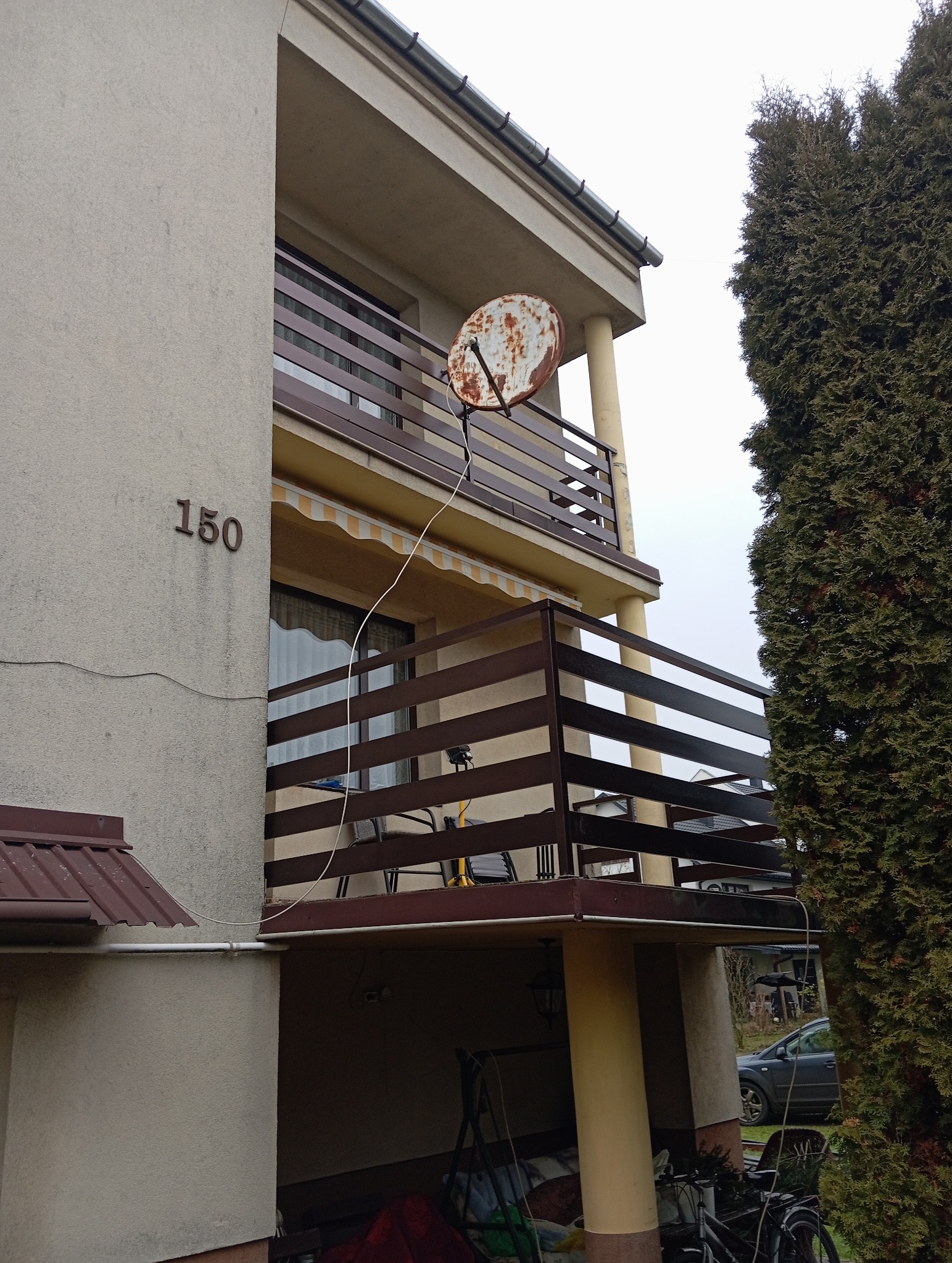 Dwukondygnacyjny budynek z brązowymi balustradami balkonowymi, zardzewiałą anteną satelitarną na górnym balkonie i numerem domu '150' na ścianie.