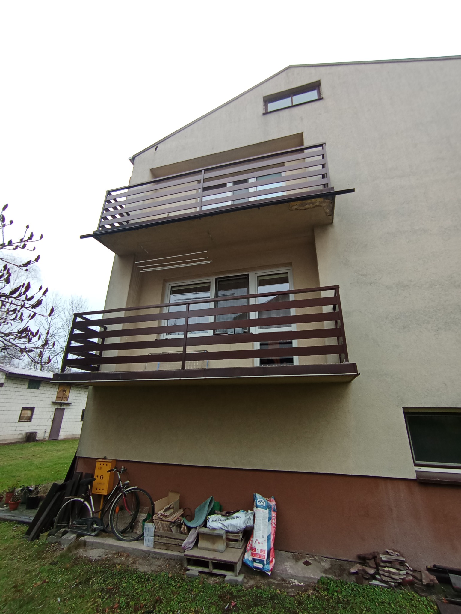 Dwu-kondygnacyjny budynek z balkonami o brązowych, poziomych balustradach; na parterze rower, skrzynki i worki z ziemią.